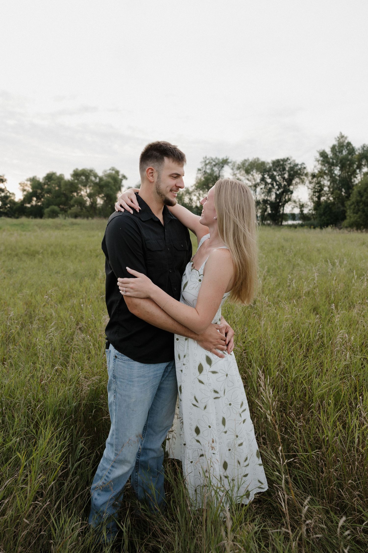 Couple embracing and smiling in tall grass field at dusk, woman in floral dress hugging man.