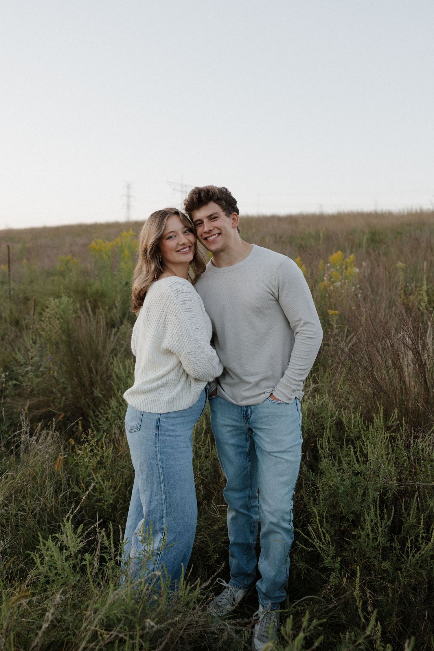 Smiling couple standing close together in a wildflower field at dusk, wearing matching cream and white sweaters.