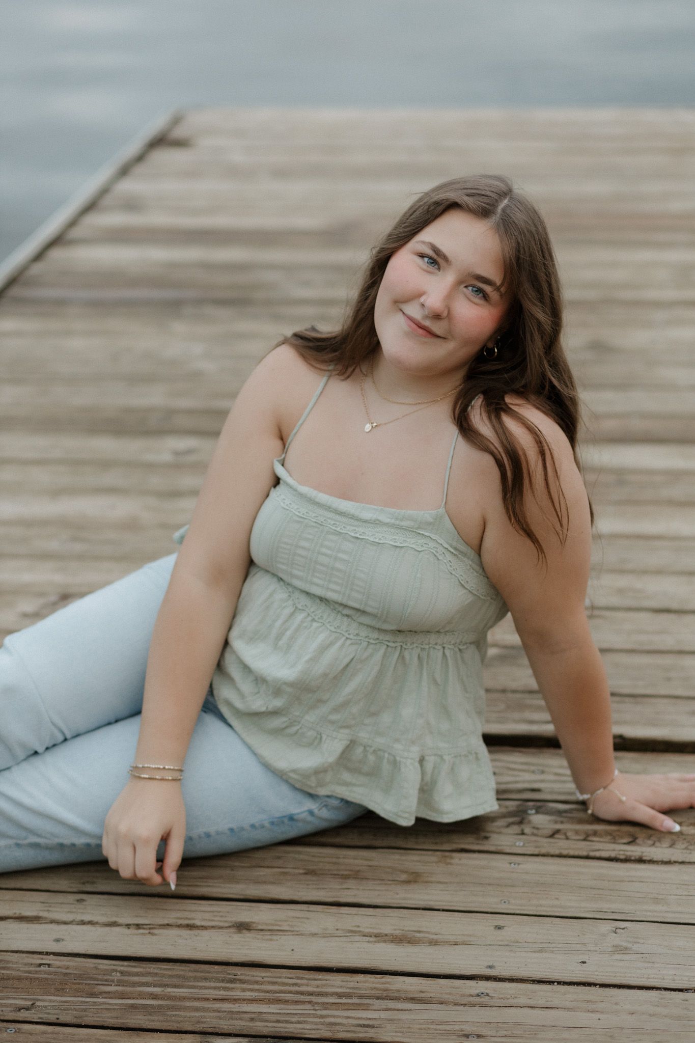 Young woman in mint top and jeans sitting on wooden dock by calm water smiling at camera.