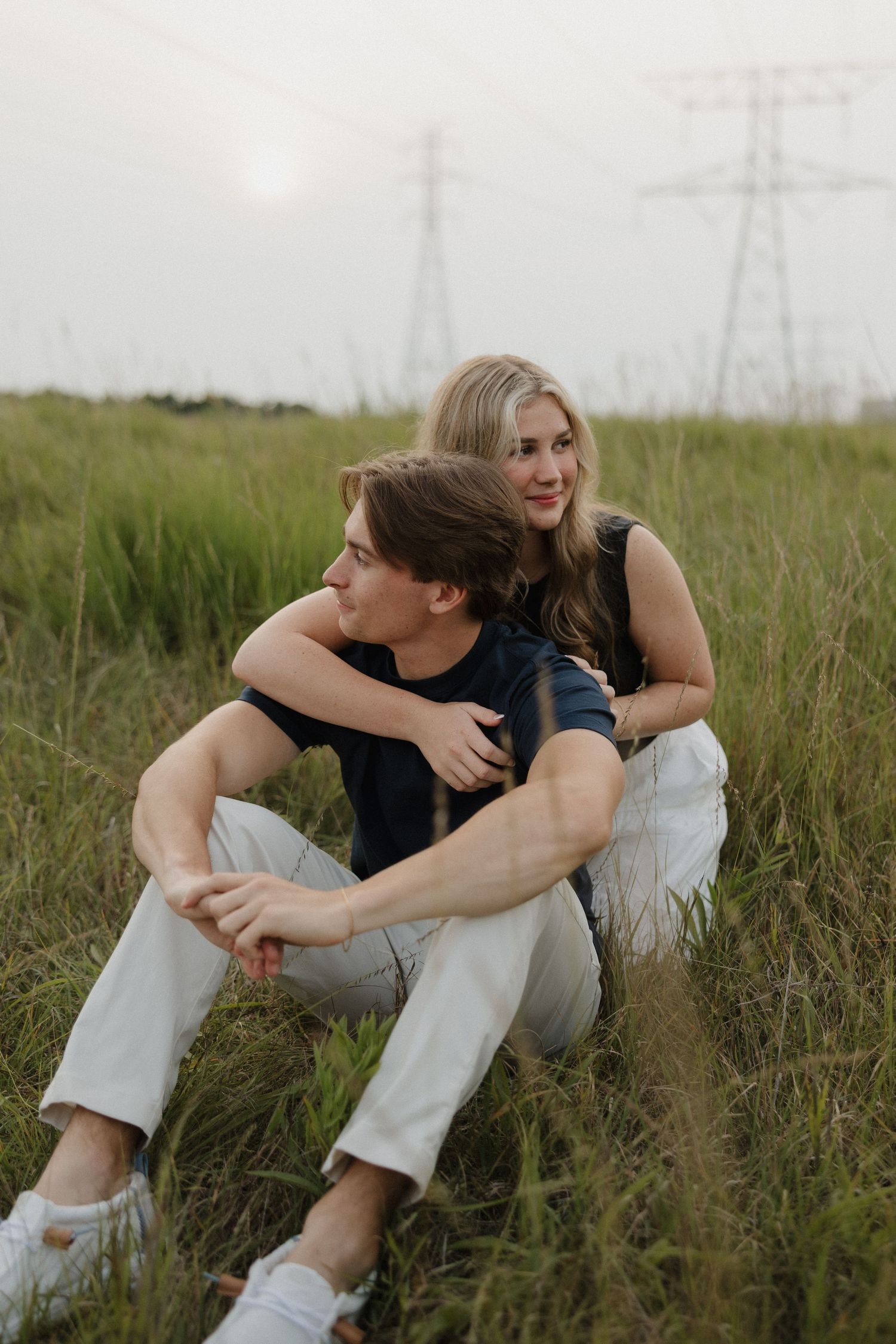 Young couple sitting in green field, woman embracing man from behind near power lines at sunset.