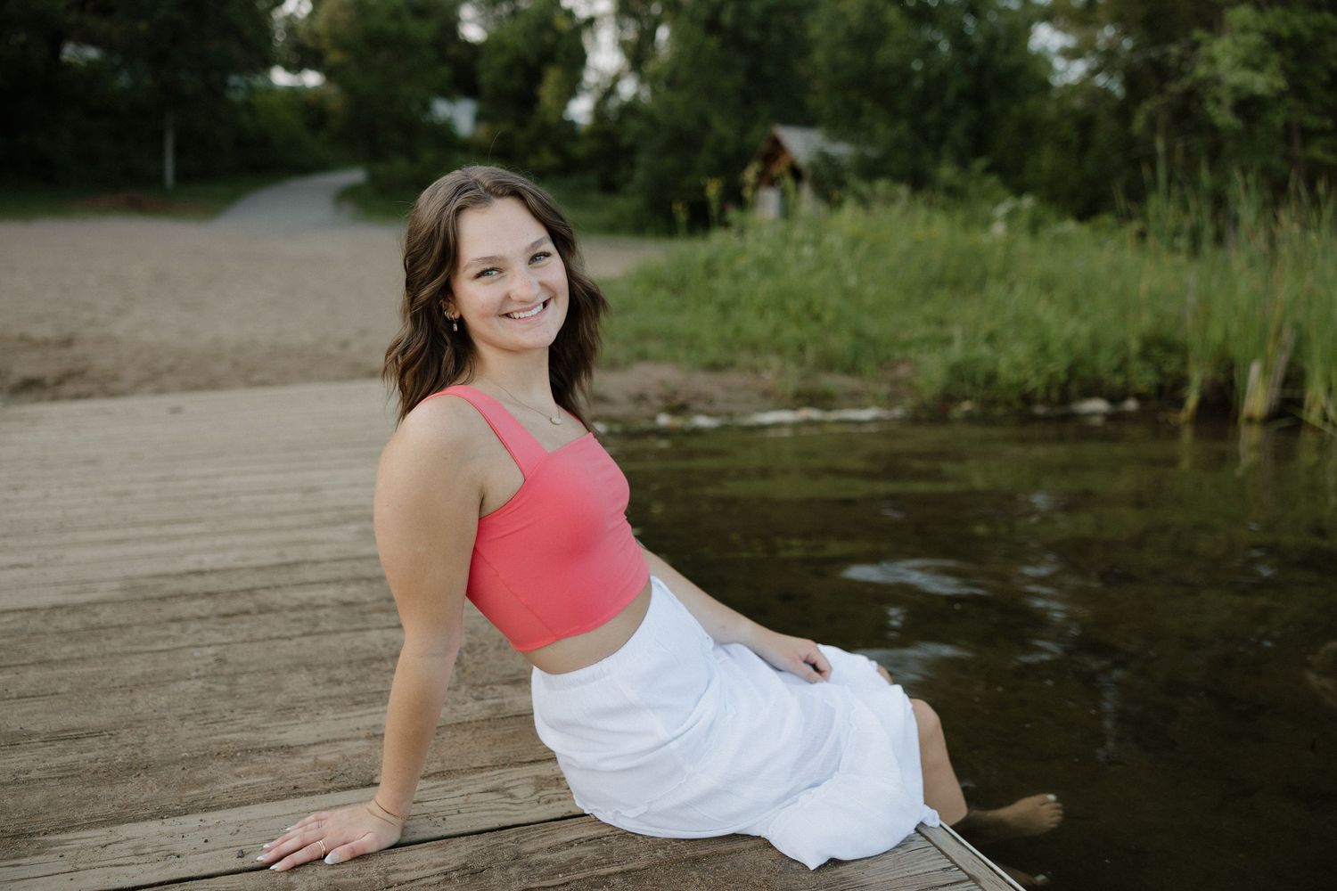 Smiling young woman in a coral top and white skirt sitting on a wooden dock by a river.