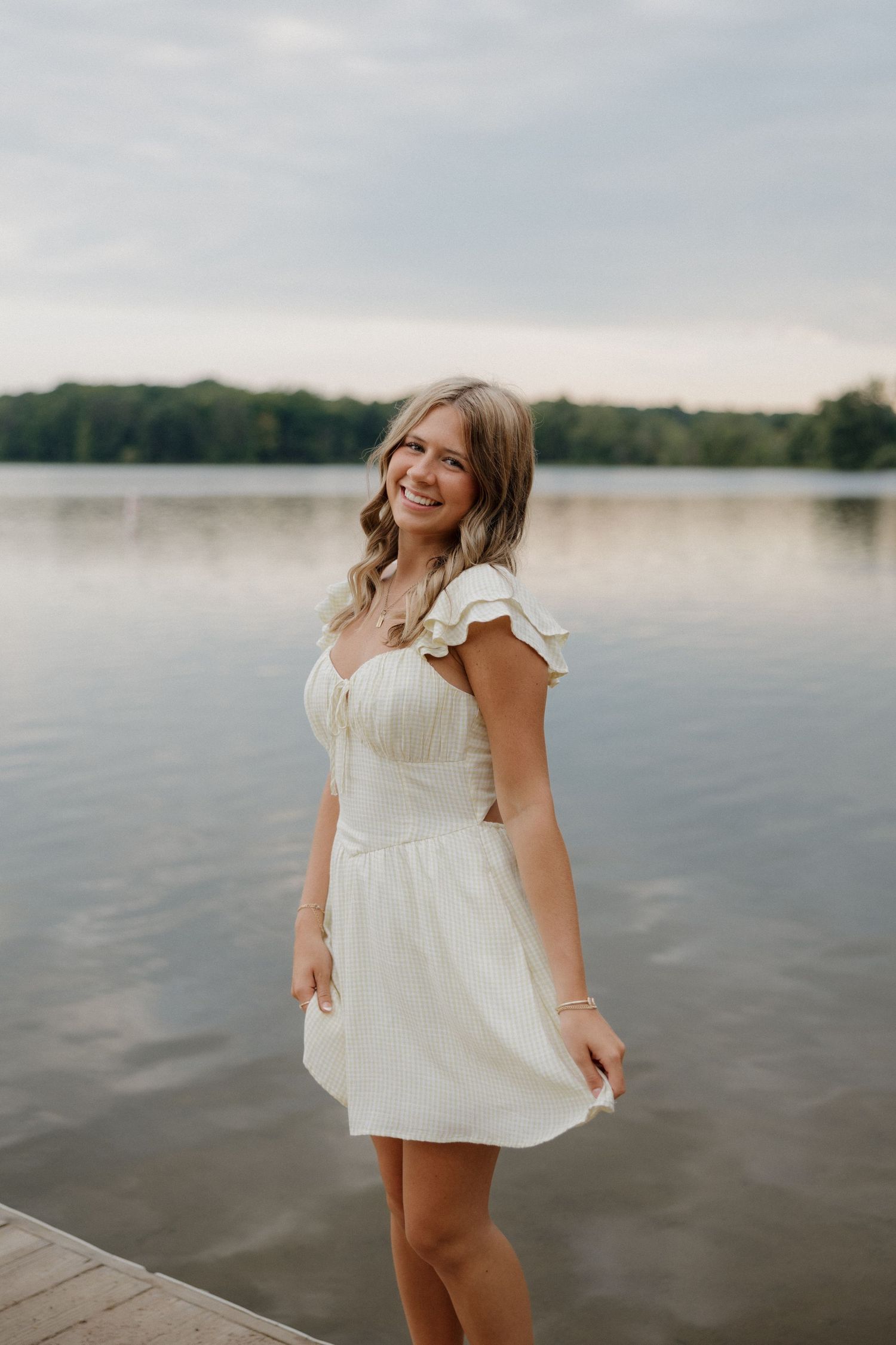 Smiling blonde woman in white flutter-sleeve dress stands on dock with calm lake behind her.