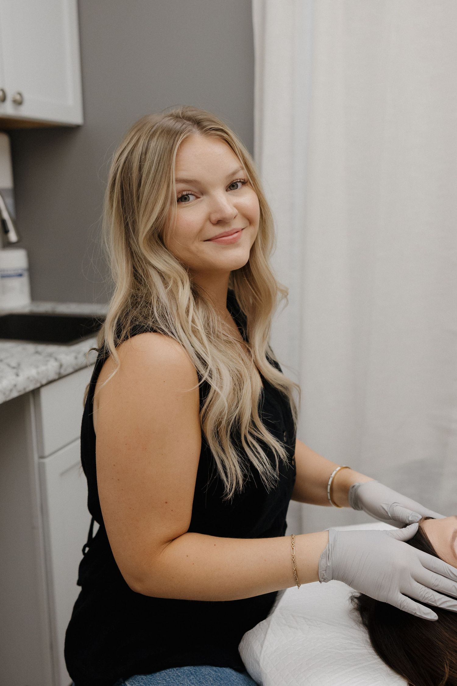 Blonde esthetician in black top and gloves smiling while performing a facial treatment on client.