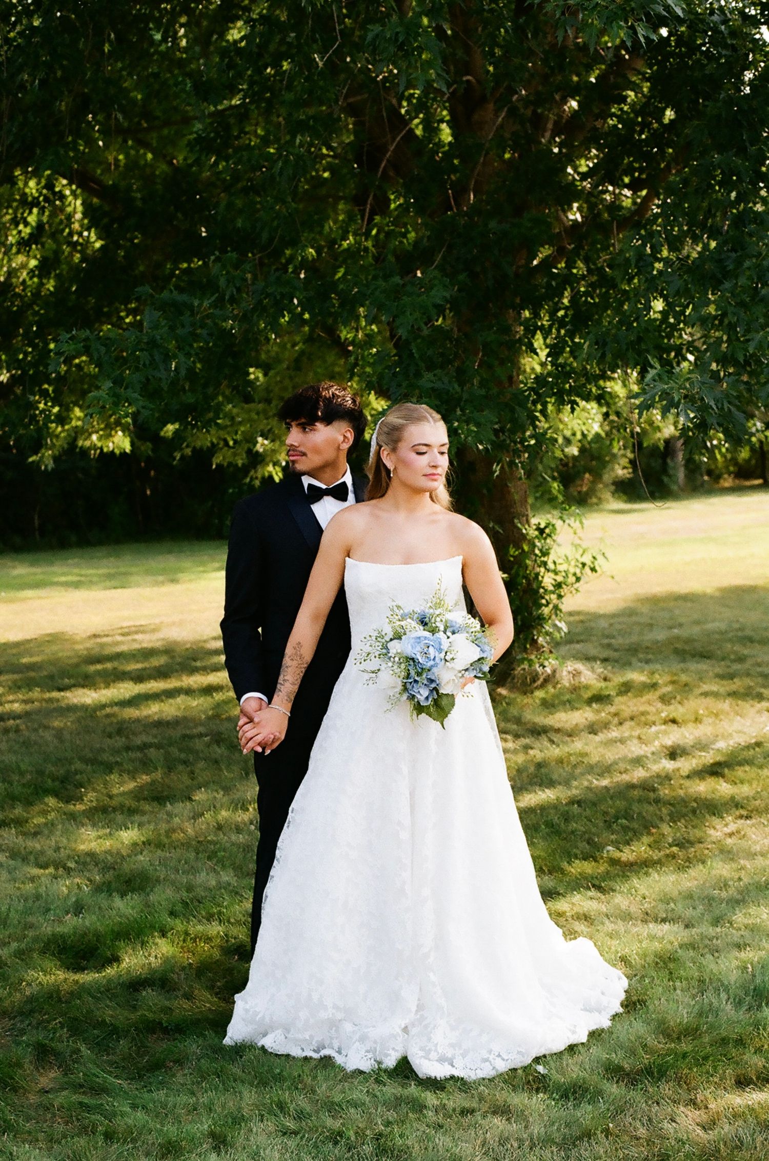 Bride in strapless lace gown holding blue bouquet stands with groom in black tuxedo outdoors near green trees.
