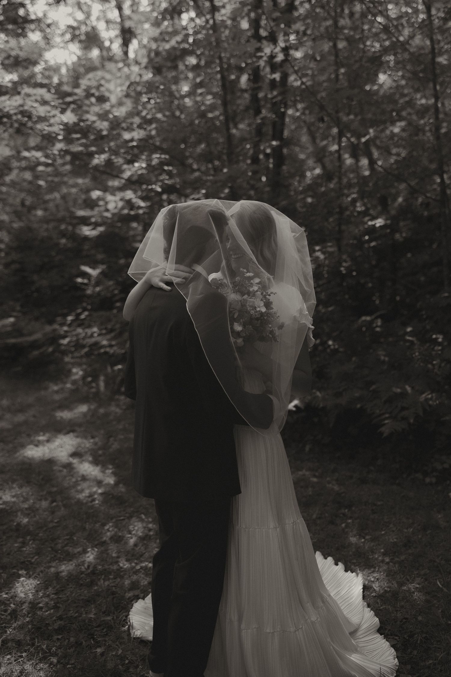 Black and white photo of bride and groom kissing under a veil in a lush forest setting.