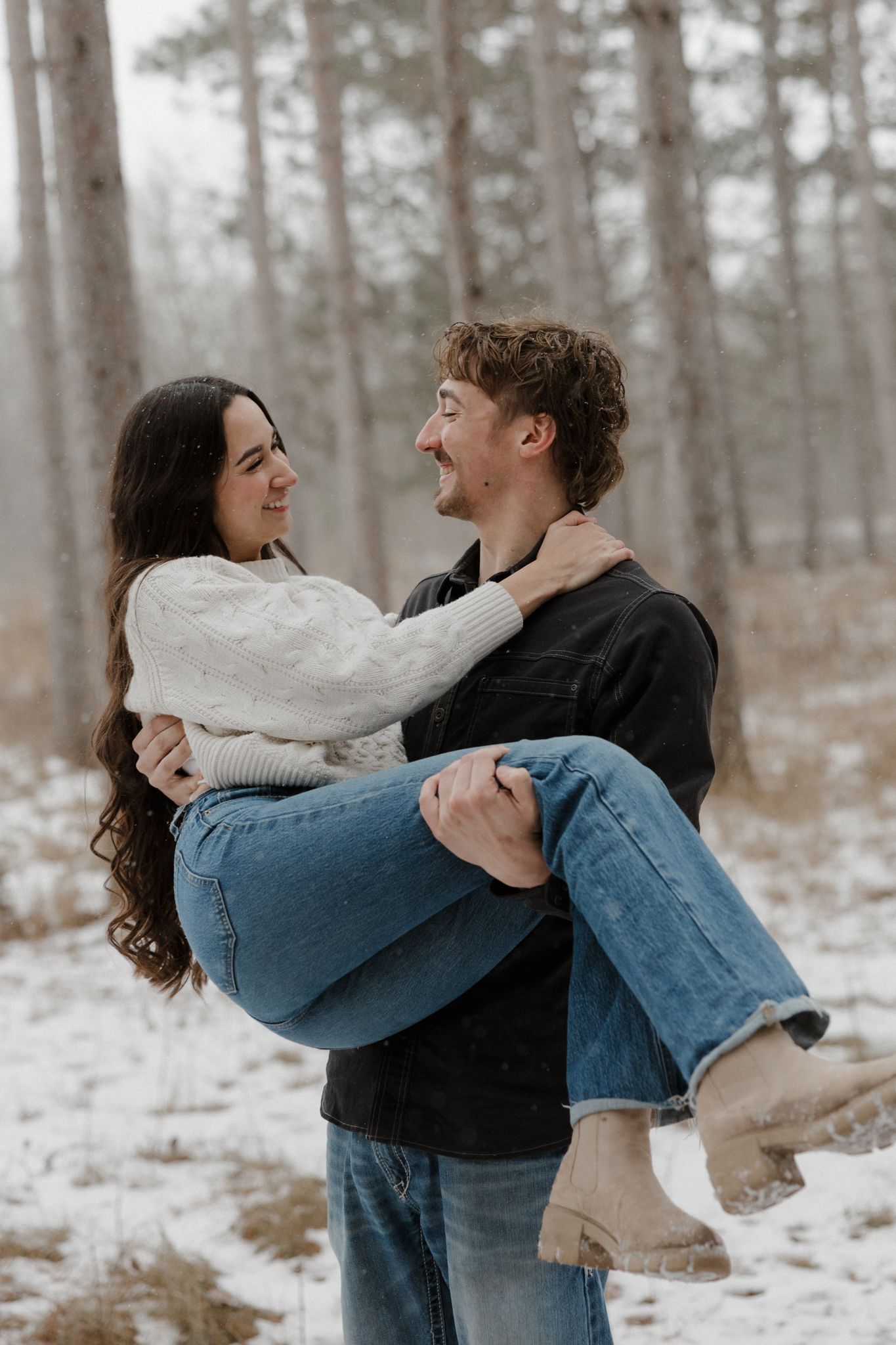 Man carrying woman in his arms in snowy forest, couple smiling at each other romantically.