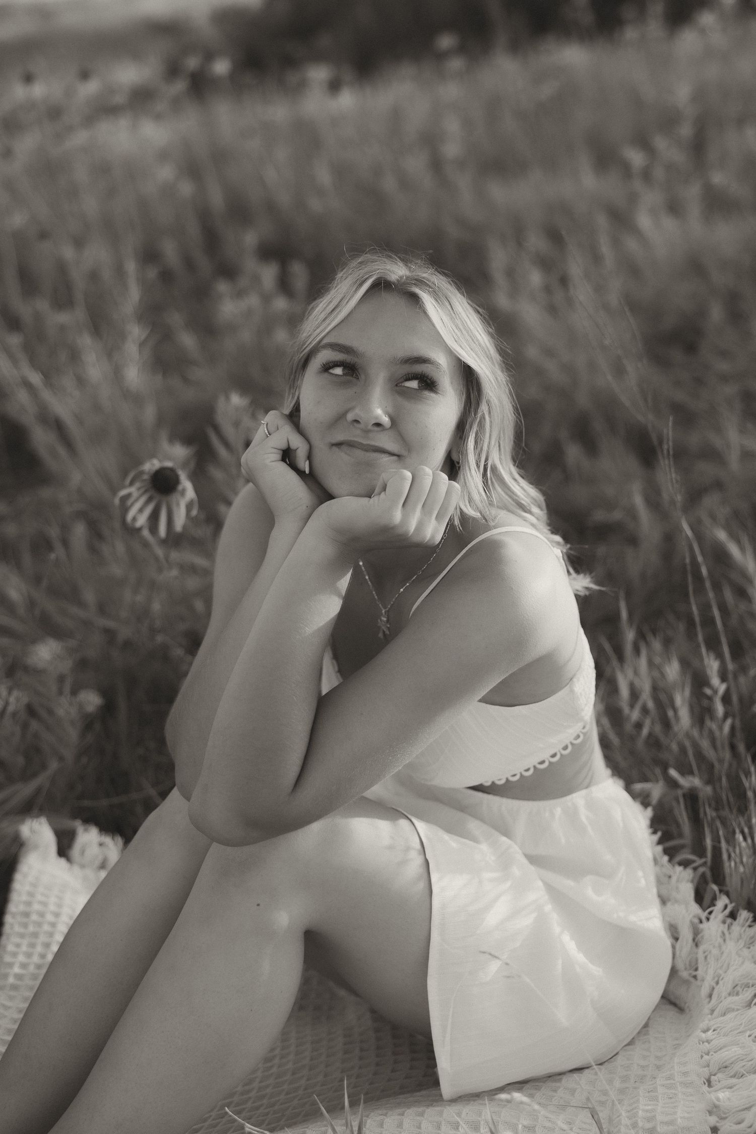 Black and white photo of blonde woman sitting in field holding daisy flower.