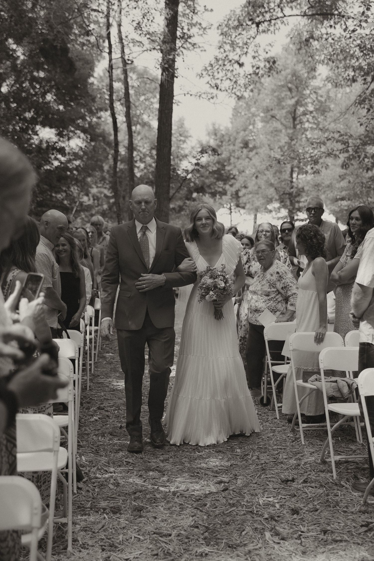 Wedding ceremony processional in a wooded outdoor setting with white chairs lining a natural aisle.