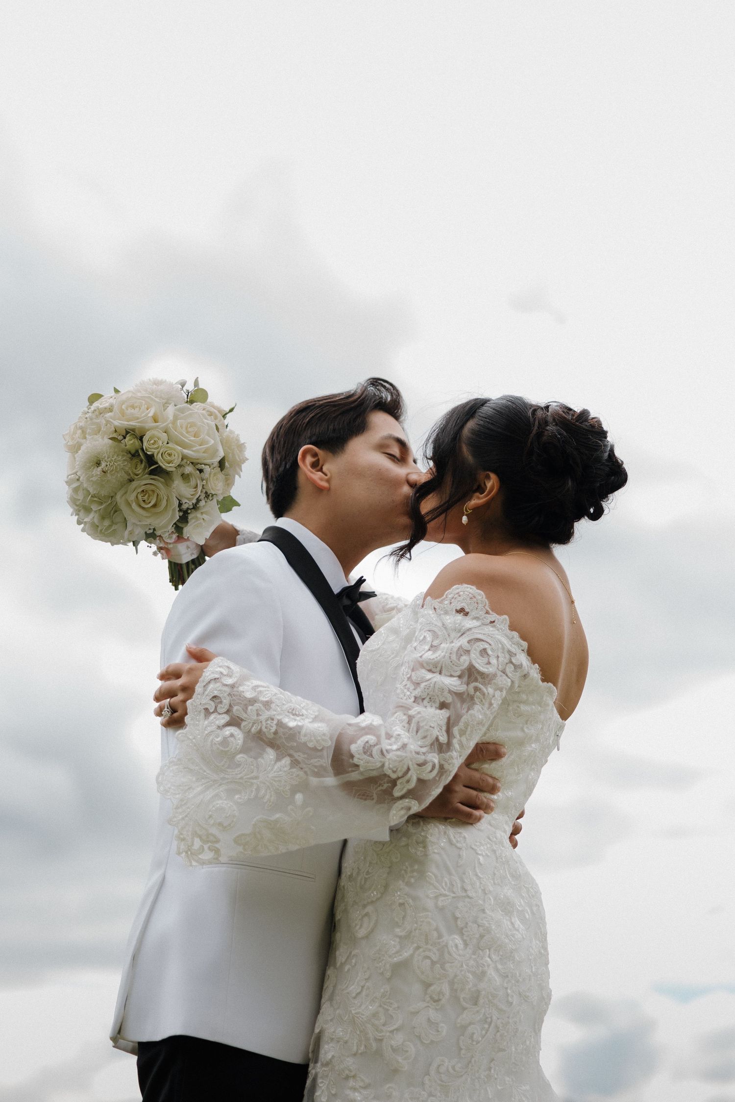 A wedding couple embraces while holding a white rose bouquet against a cloudy sky backdrop.