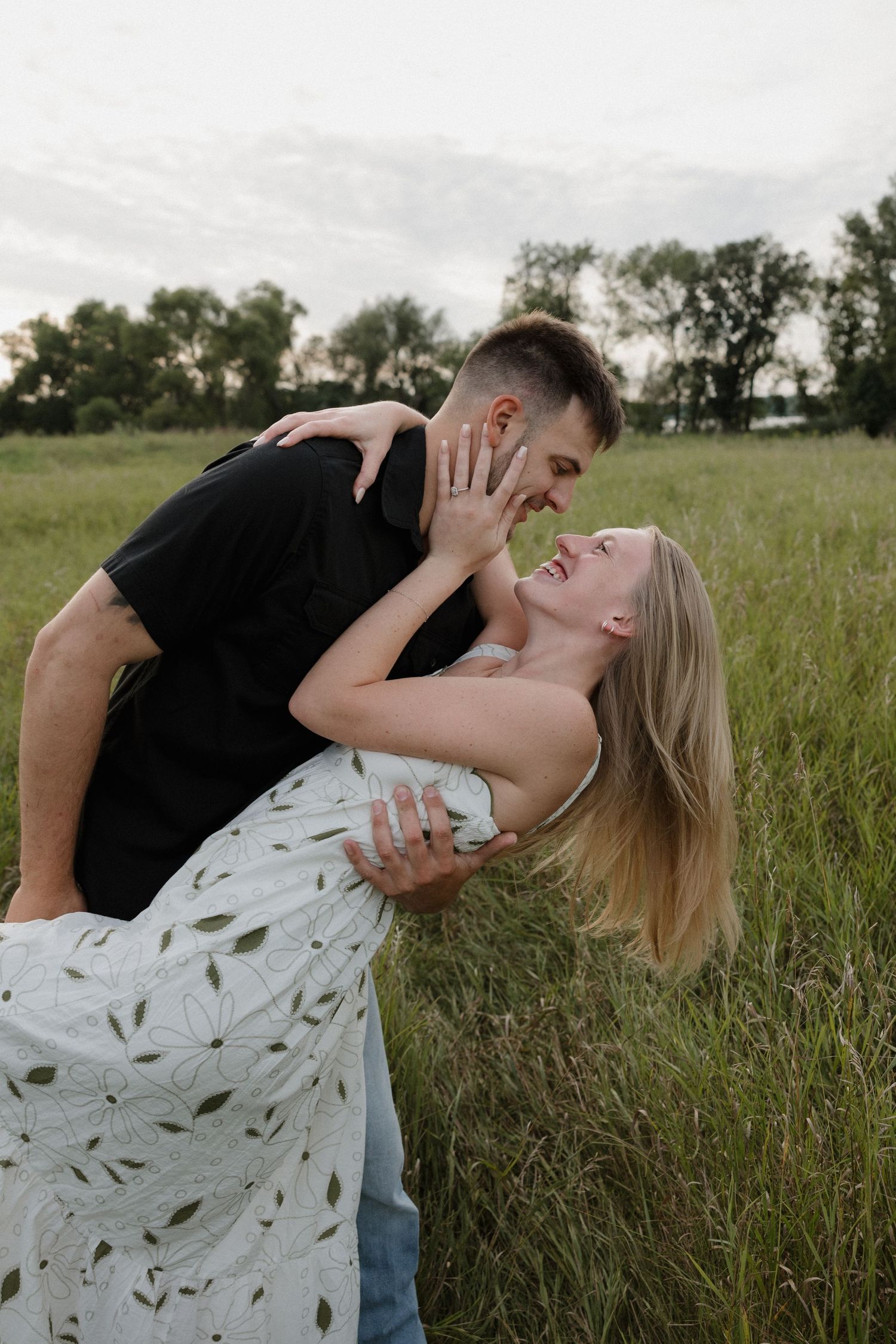 Couple sharing a romantic dip embrace while laughing together in a lush green field at dusk.