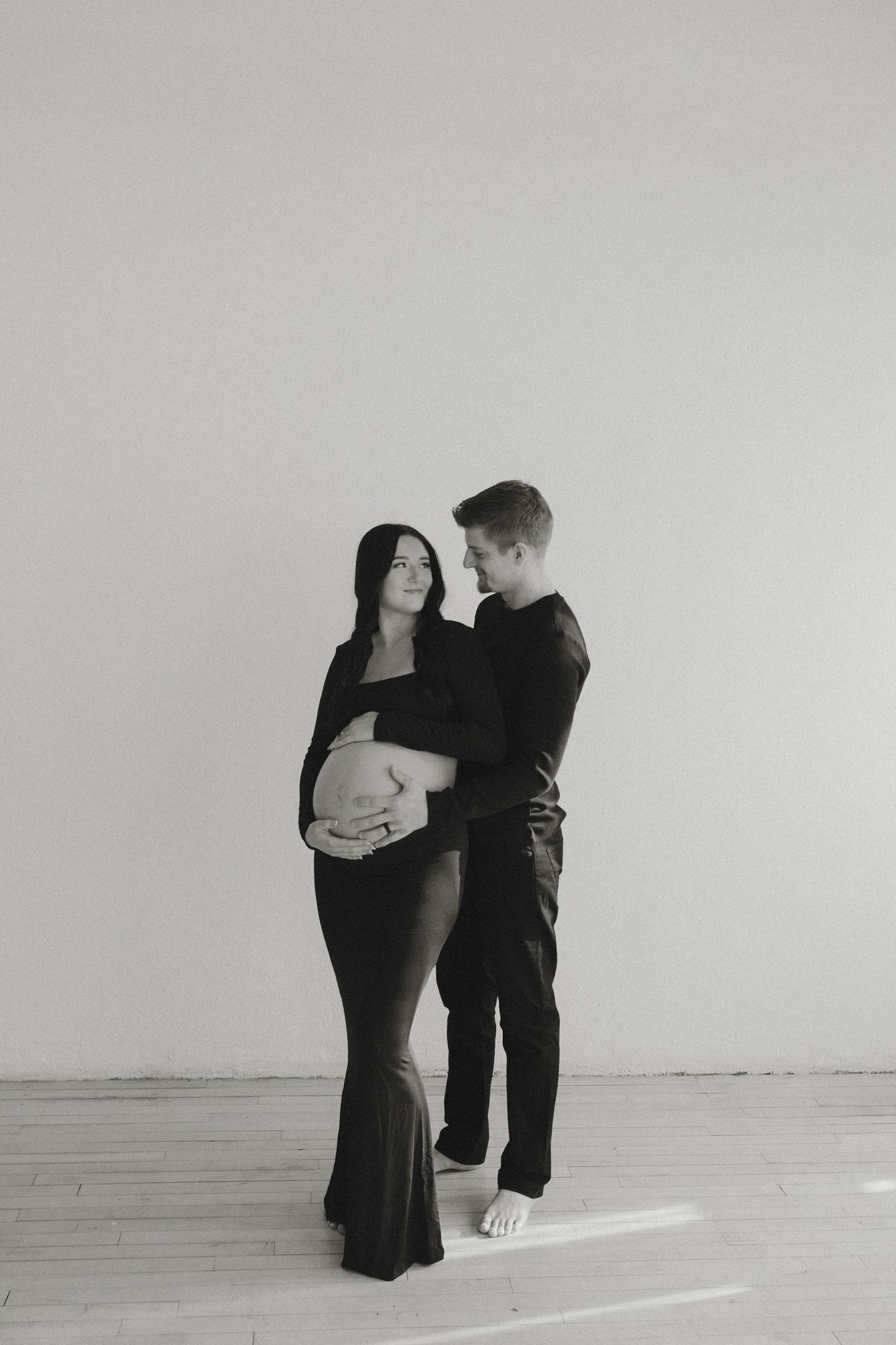 Couple embracing during black and white maternity photoshoot in minimal white studio.