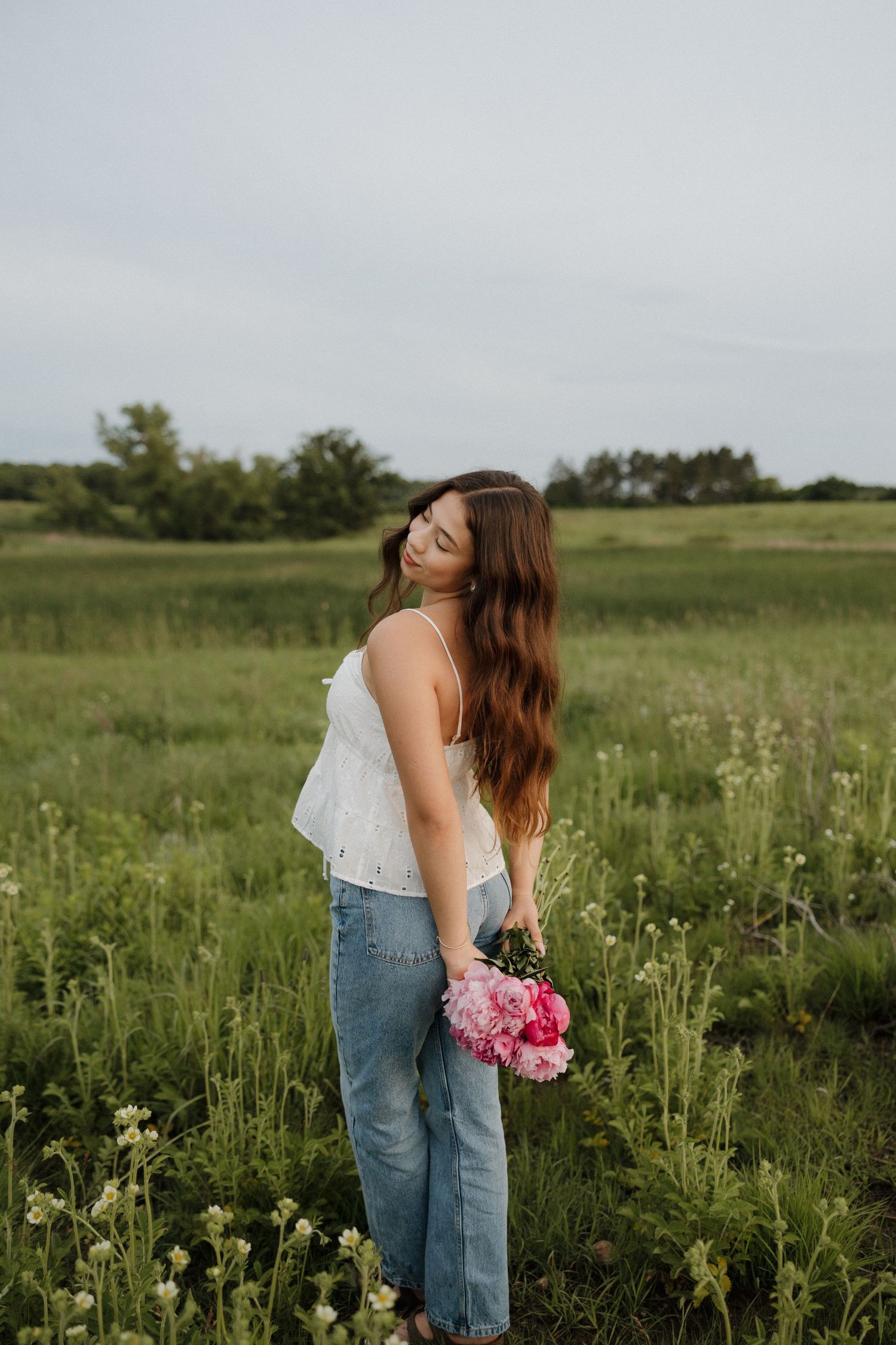 Young woman in white camisole and jeans holding pink peonies while standing in a wildflower meadow.