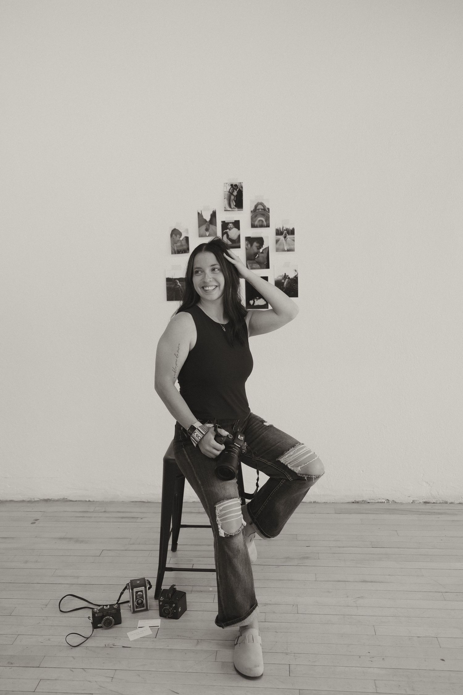 Black and white photo of smiling woman on stool holding camera with photos on wall and vintage cameras on floor.