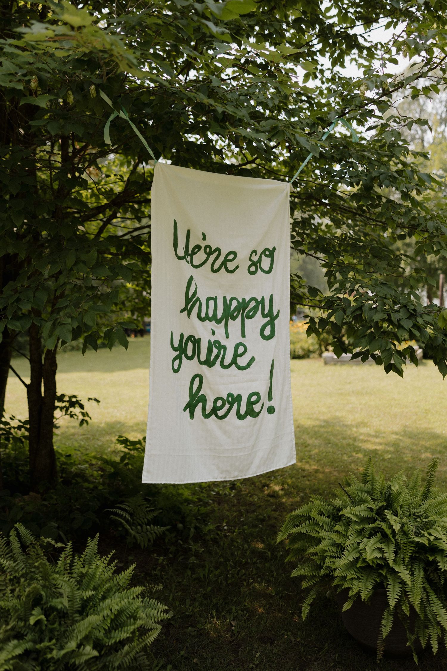 White banners with green lettering spelling 'happy you're here' hang between trees in a garden setting.