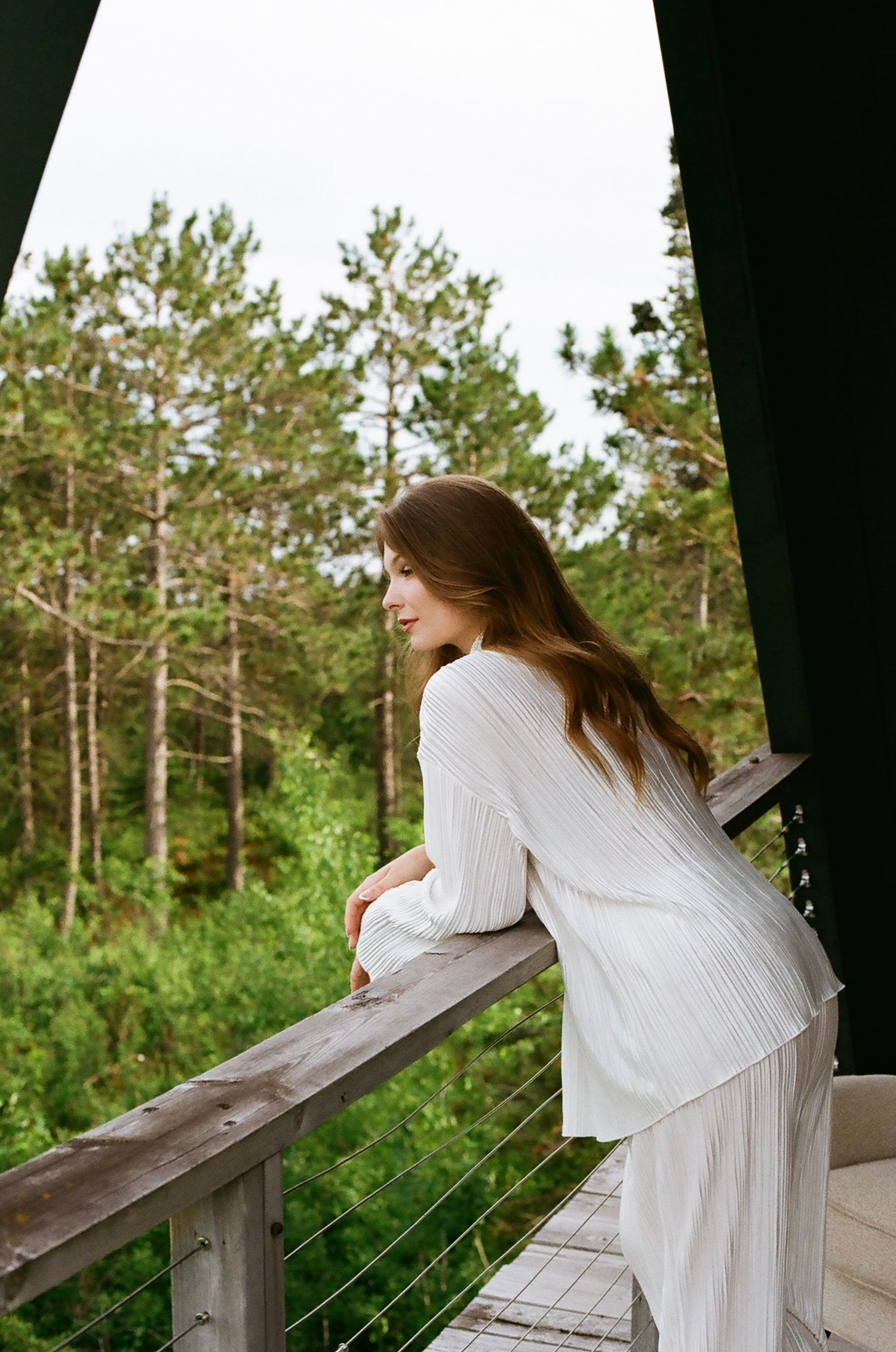 A woman in a white outfit leans on a wooden balcony railing, gazing at lush green forest trees.