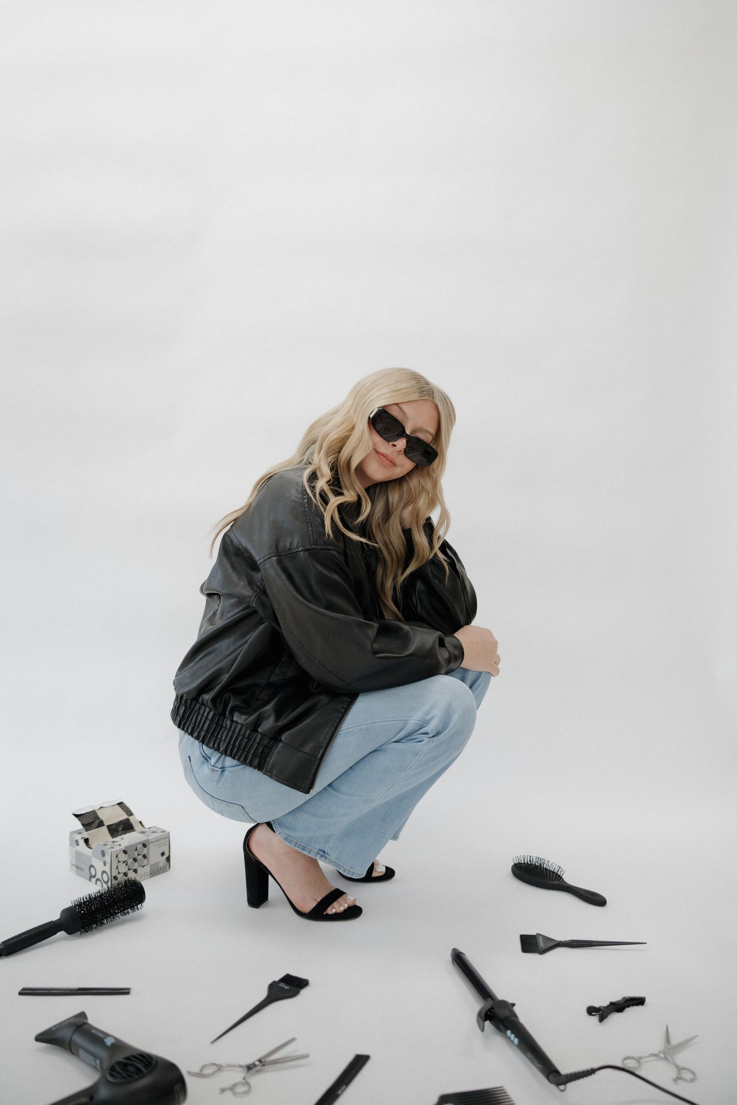 Blonde woman in leather jacket and sunglasses crouching among scattered hair styling tools on white floor.