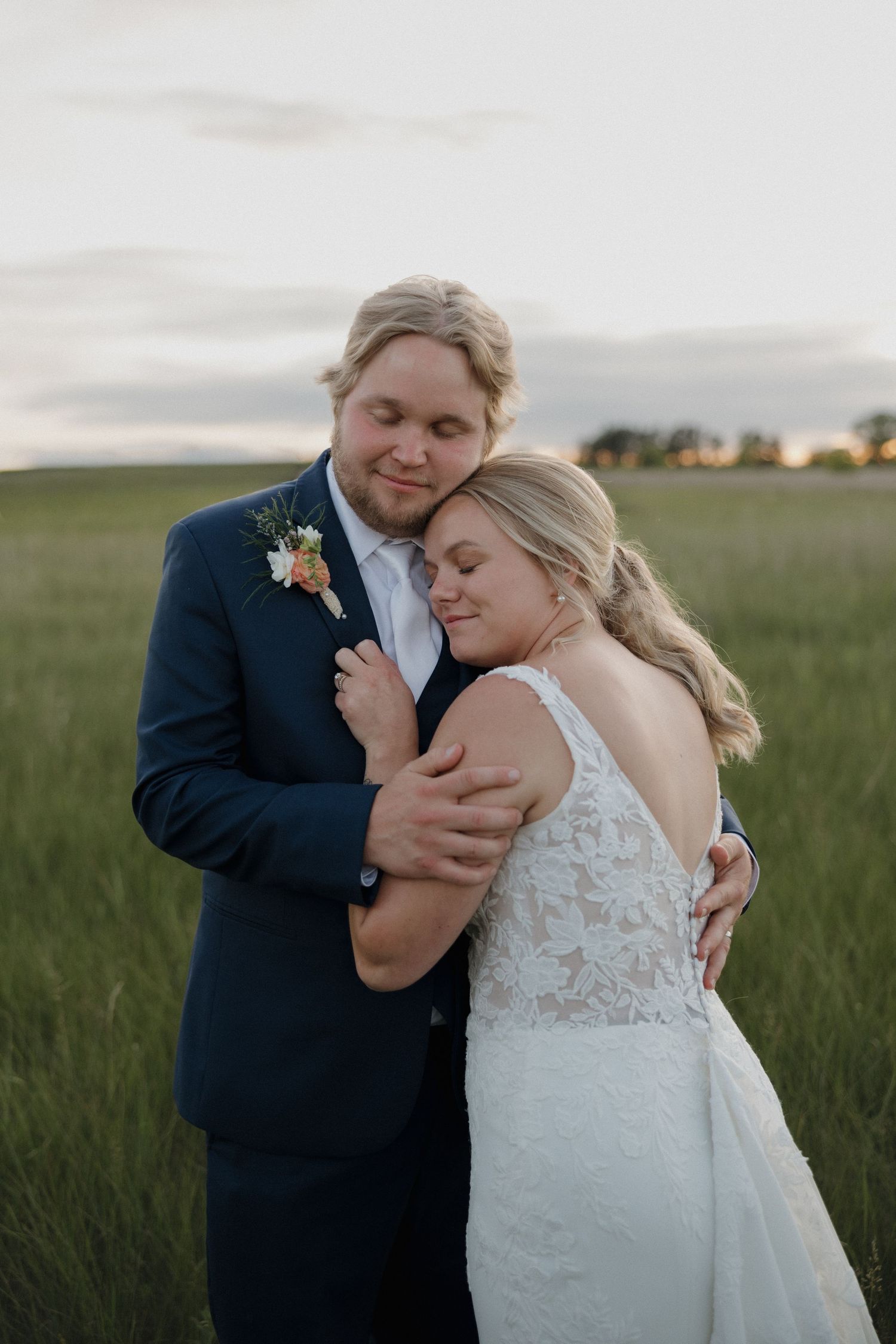 Bride and groom embracing in a green field at sunset, groom in navy suit with floral boutonniere.
