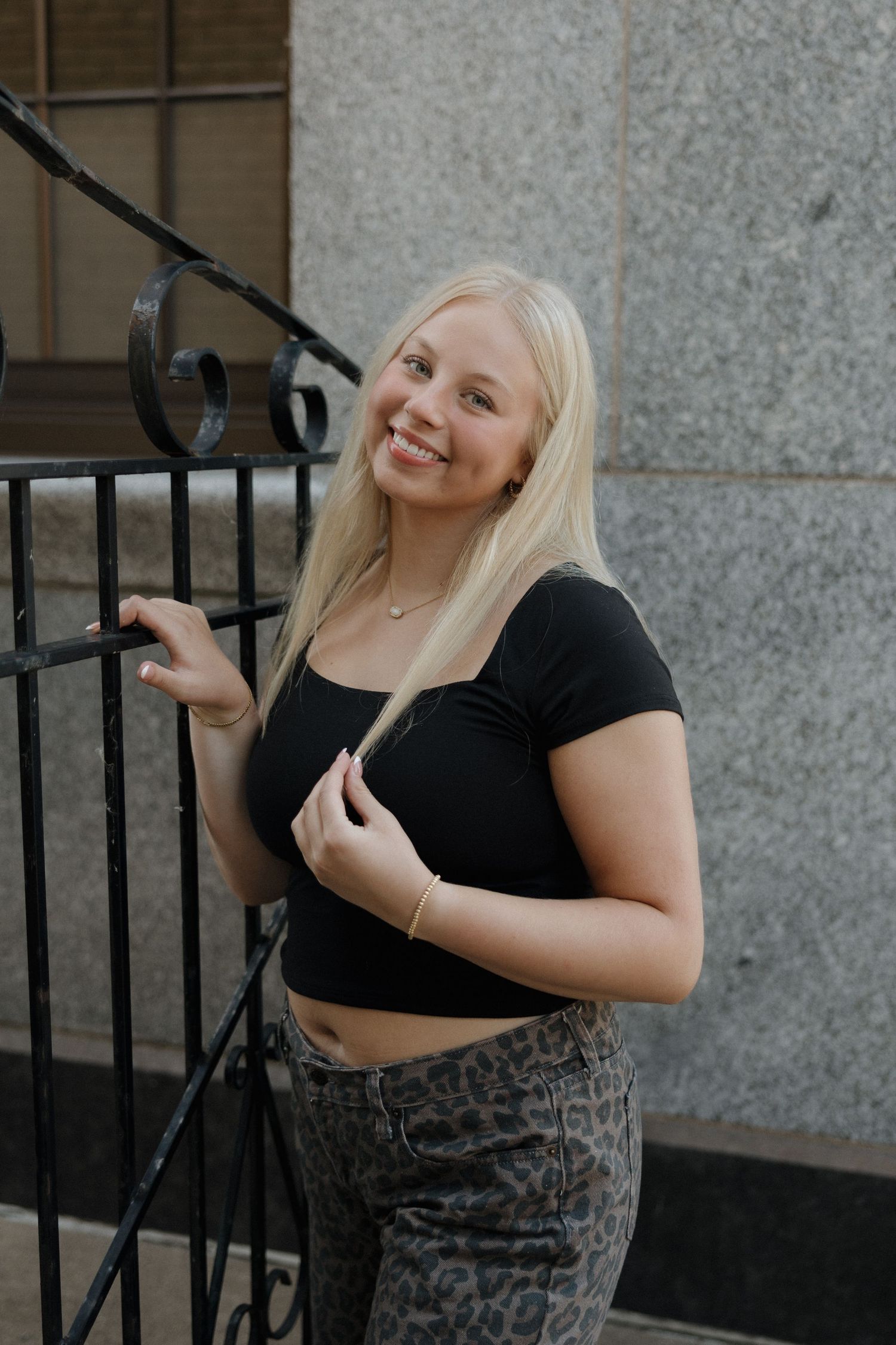 Smiling blonde woman in black crop top and leopard pants posing beside a wrought iron railing outdoors.