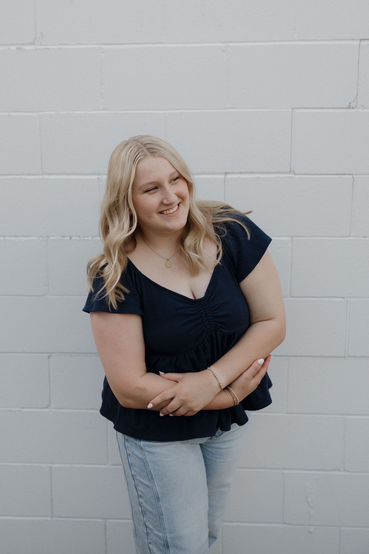 Smiling blonde woman in navy top and jeans posing against white brick wall.