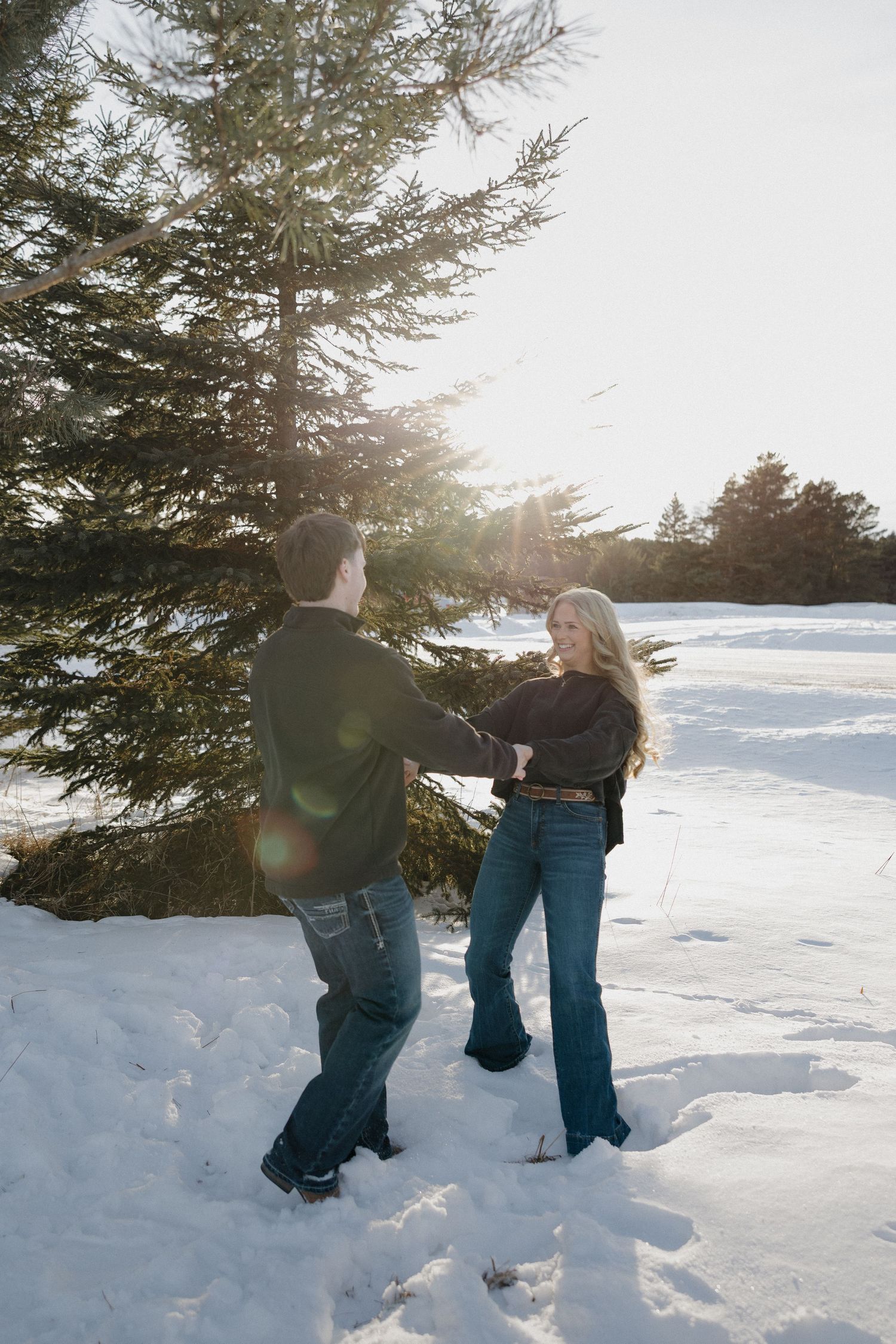 Couple holding hands and dancing playfully in the snow near a sunlit pine tree on a bright winter day.