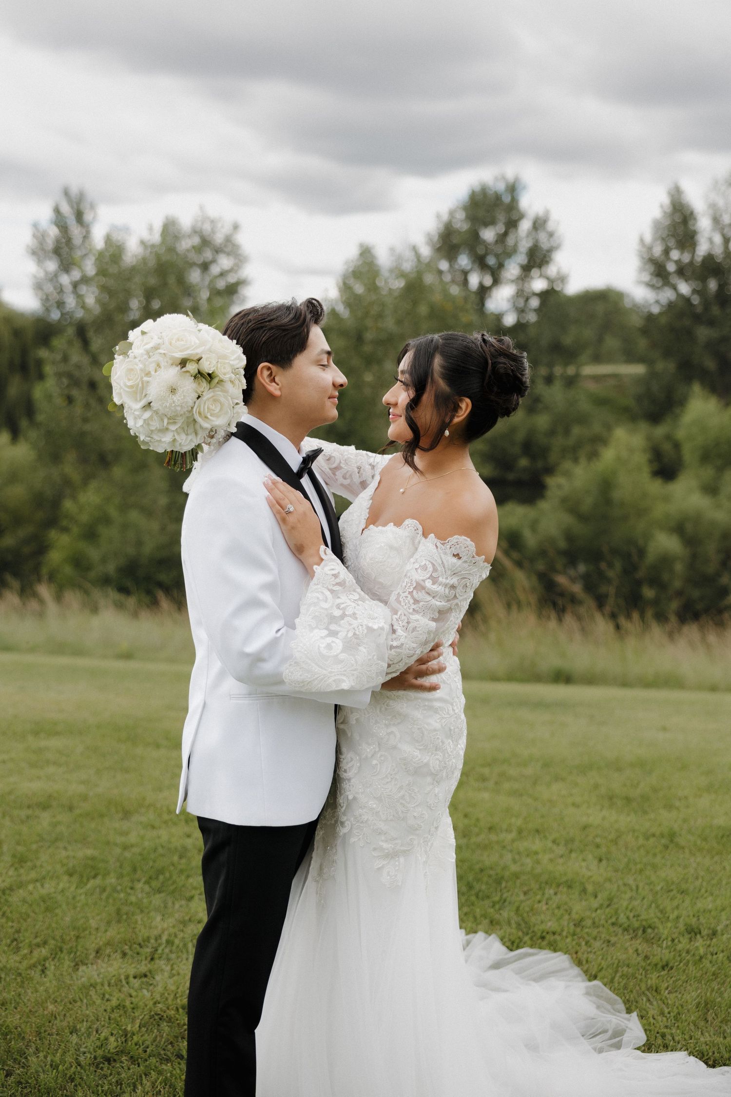 Couple in wedding attire sharing romantic moments in a grassy field under cloudy skies.