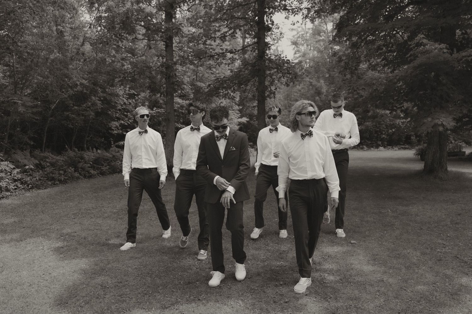 A black and white photograph shows a group of people in white shirts and dark pants walking together on a path in a wooded area.