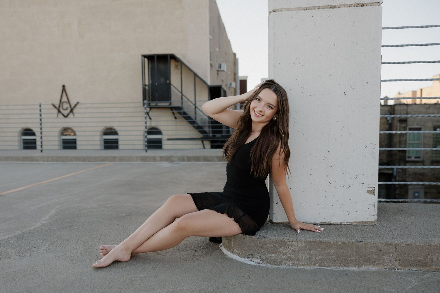 Smiling young woman in black dress sitting barefoot on a rooftop parking lot against a concrete pillar.