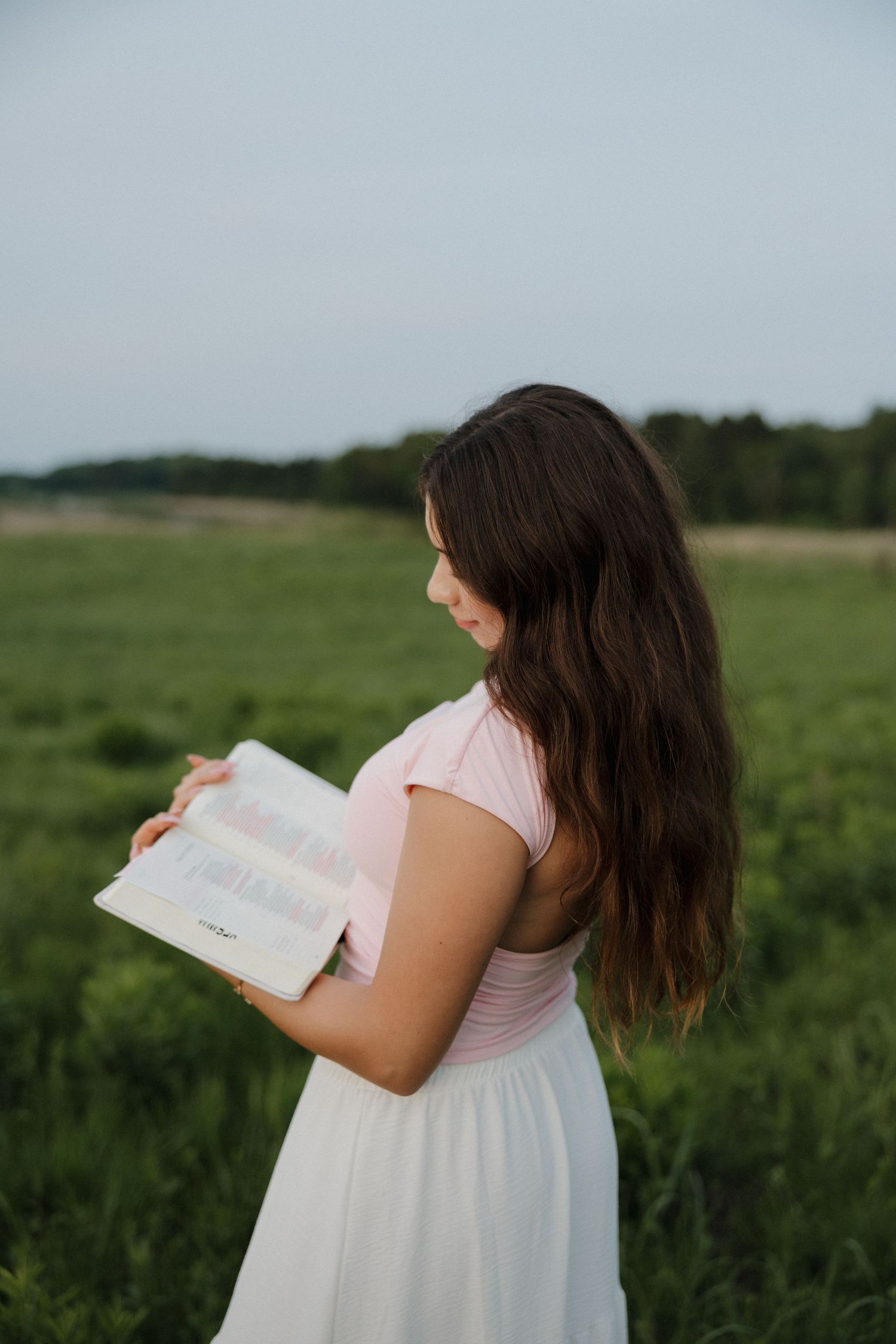 Young woman in pink top and white skirt reading an open book outdoors in a lush green field.