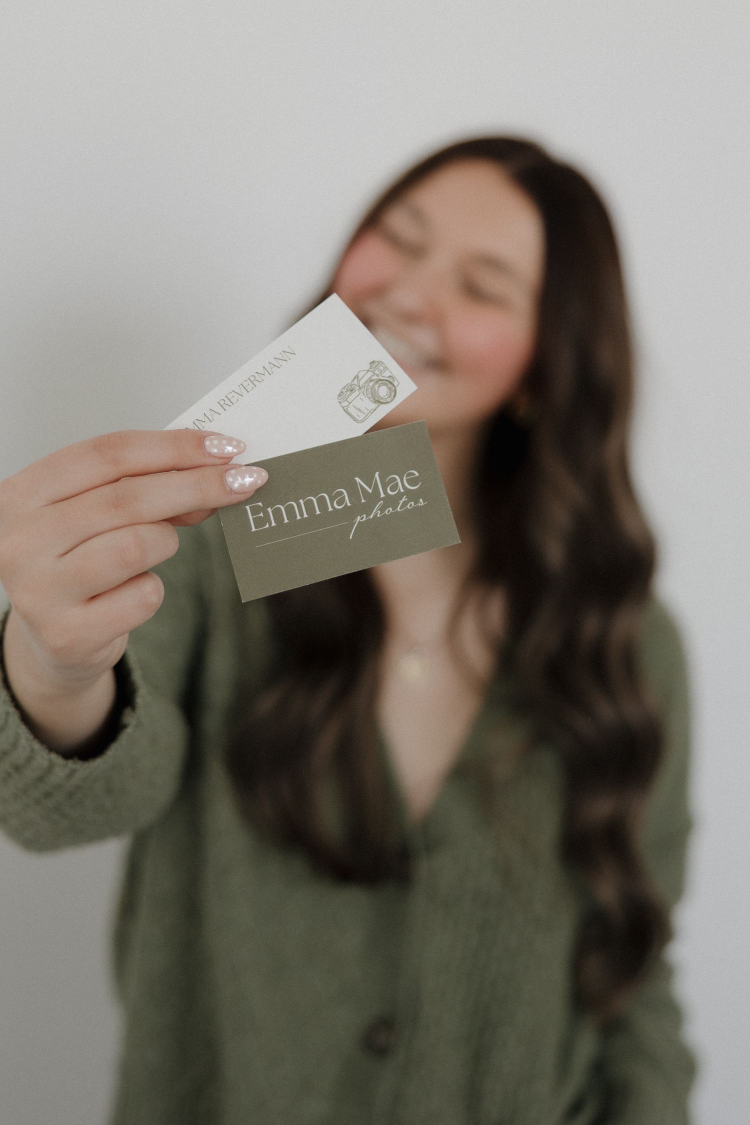 Smiling brunette woman in green sweater holding Emma Mae Photos business card toward camera.