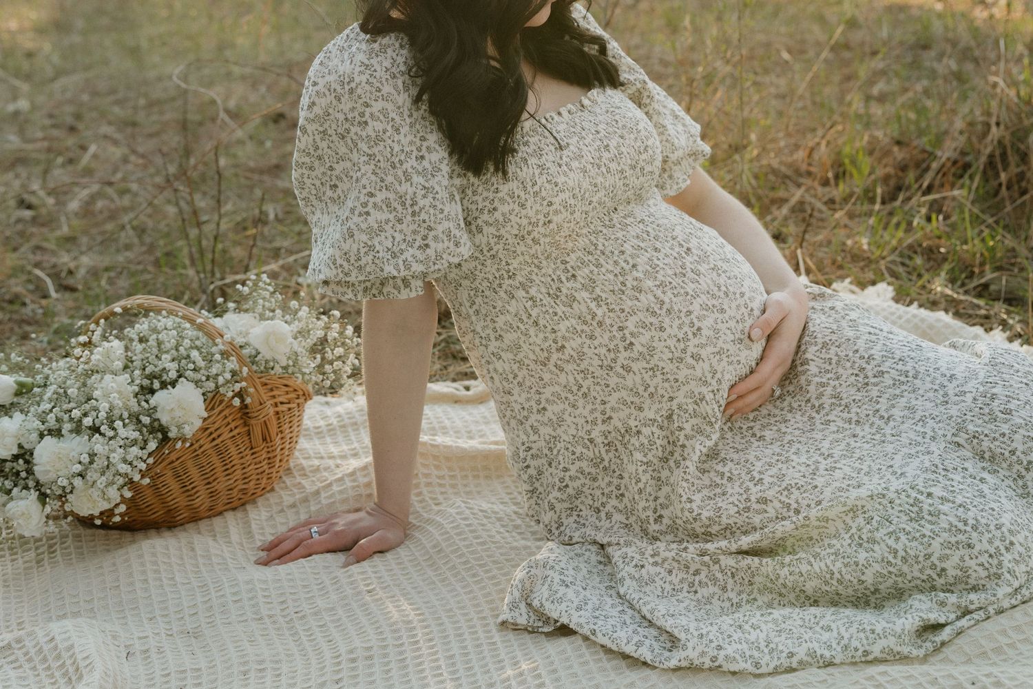 Pregnant woman in floral dress sitting on blanket outdoors with flower basket beside her.