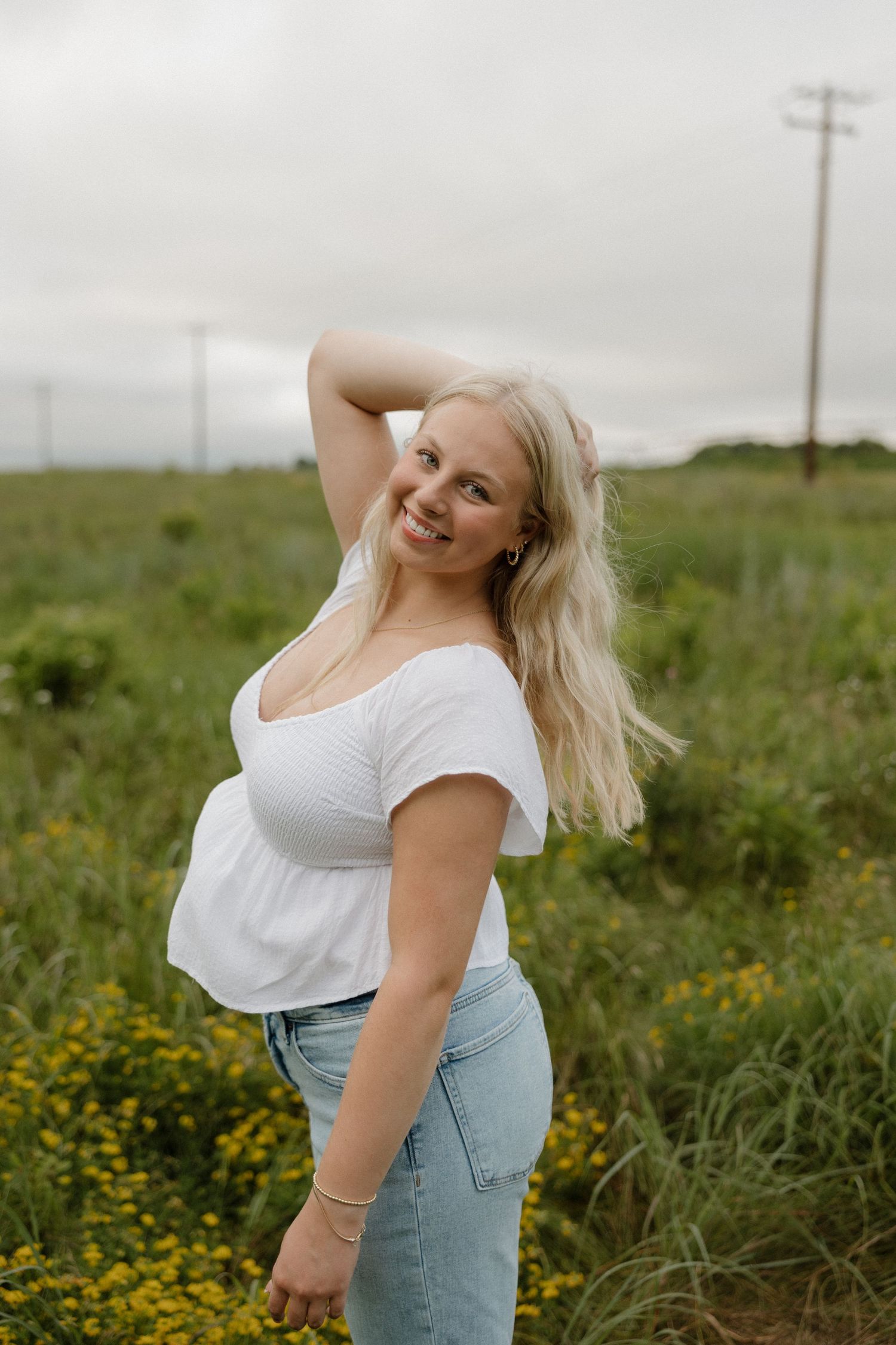 Smiling blonde woman in white top and jeans posing in a green field with yellow wildflowers on overcast day.