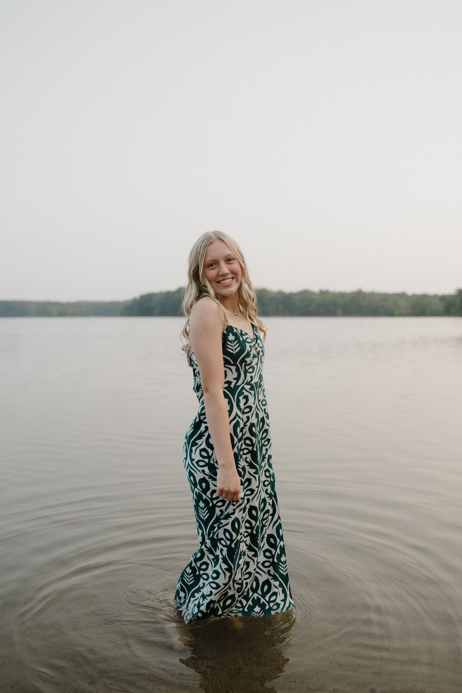 Smiling blonde woman in green patterned maxi dress standing in shallow lake water with distant treeline behind.