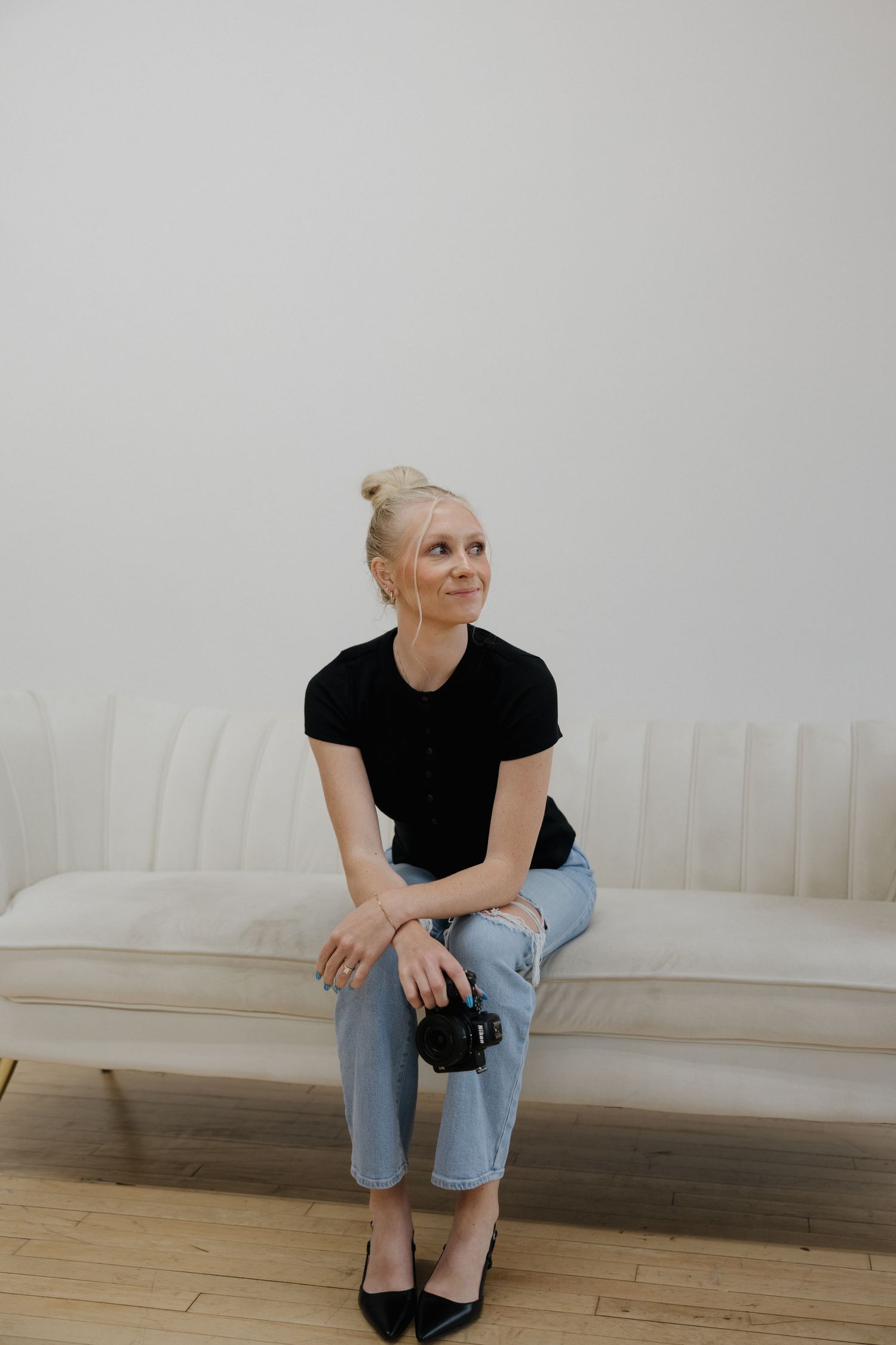 Blonde female photographer sitting on white couch holding a DSLR camera, looking up thoughtfully.