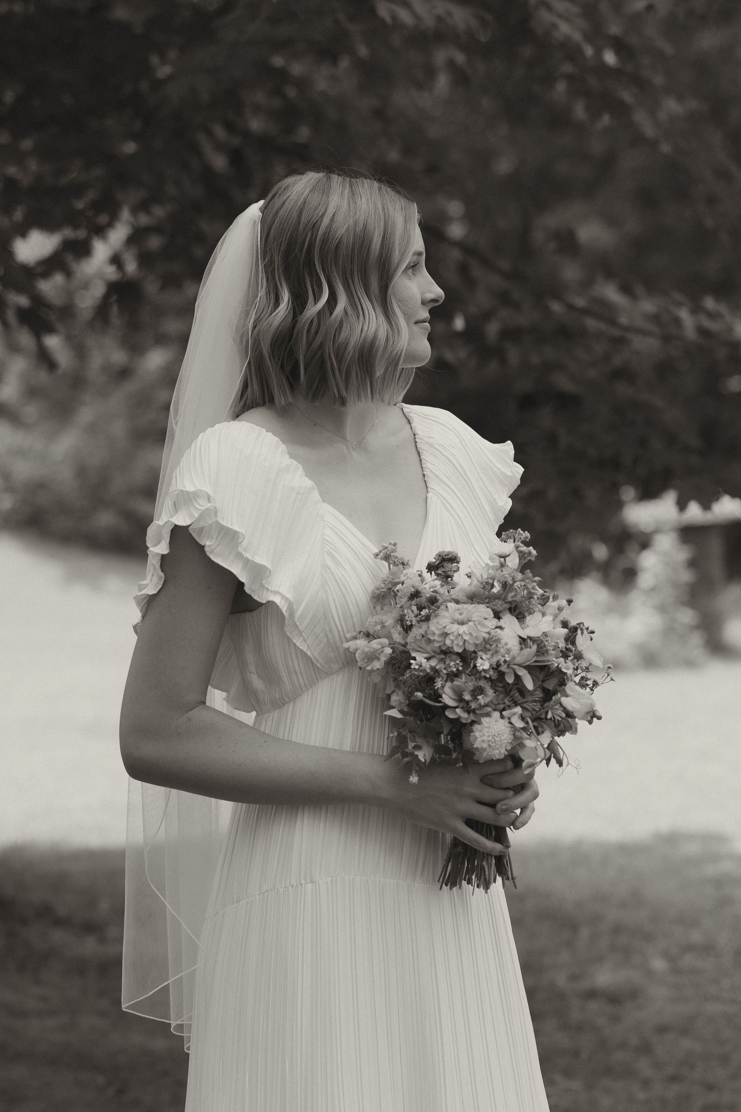 A bride in a white wedding dress holds a delicate bouquet against a natural outdoor backdrop.