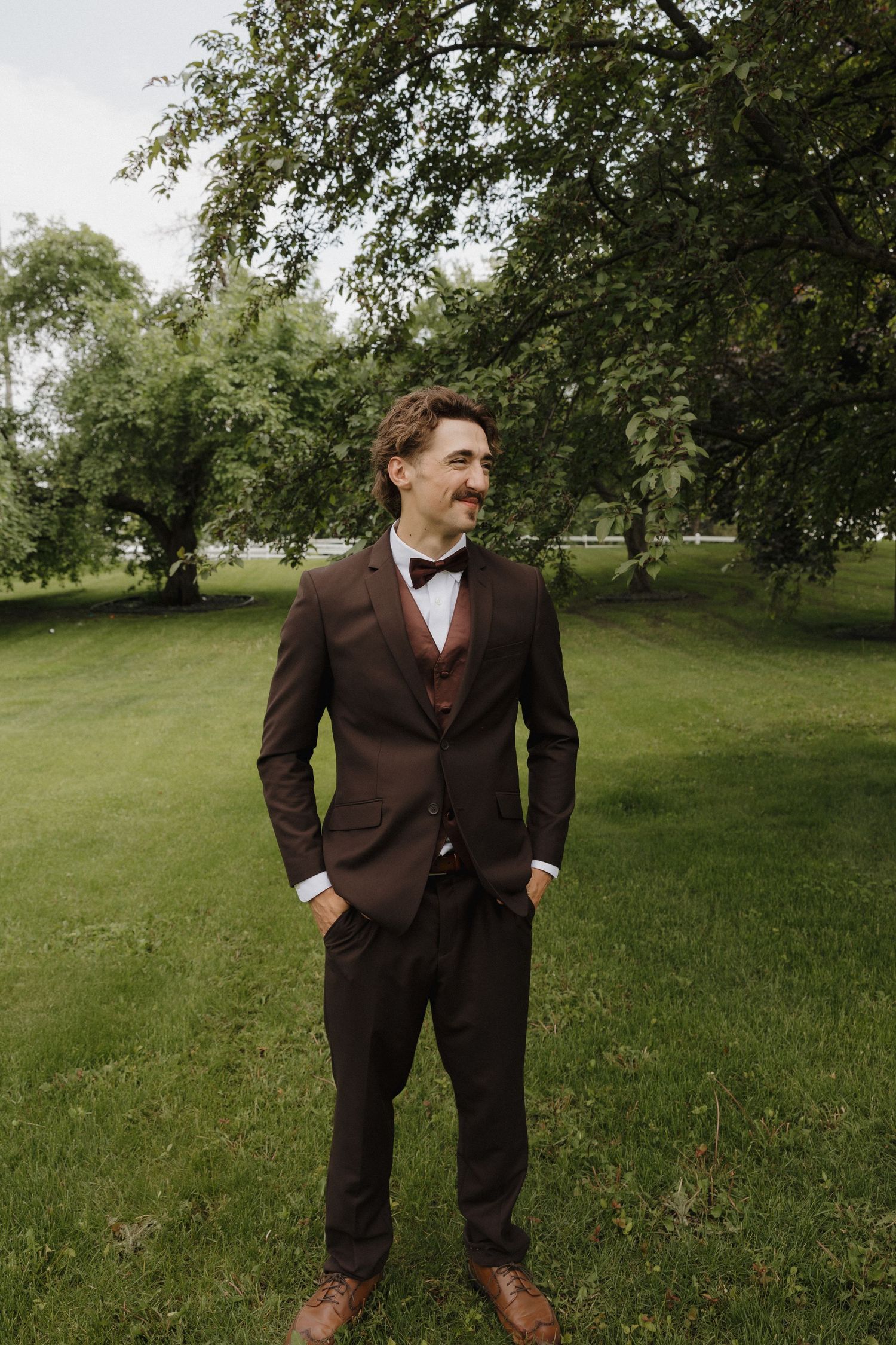 Person in a brown suit poses playfully in a green grassy field on a cloudy day.