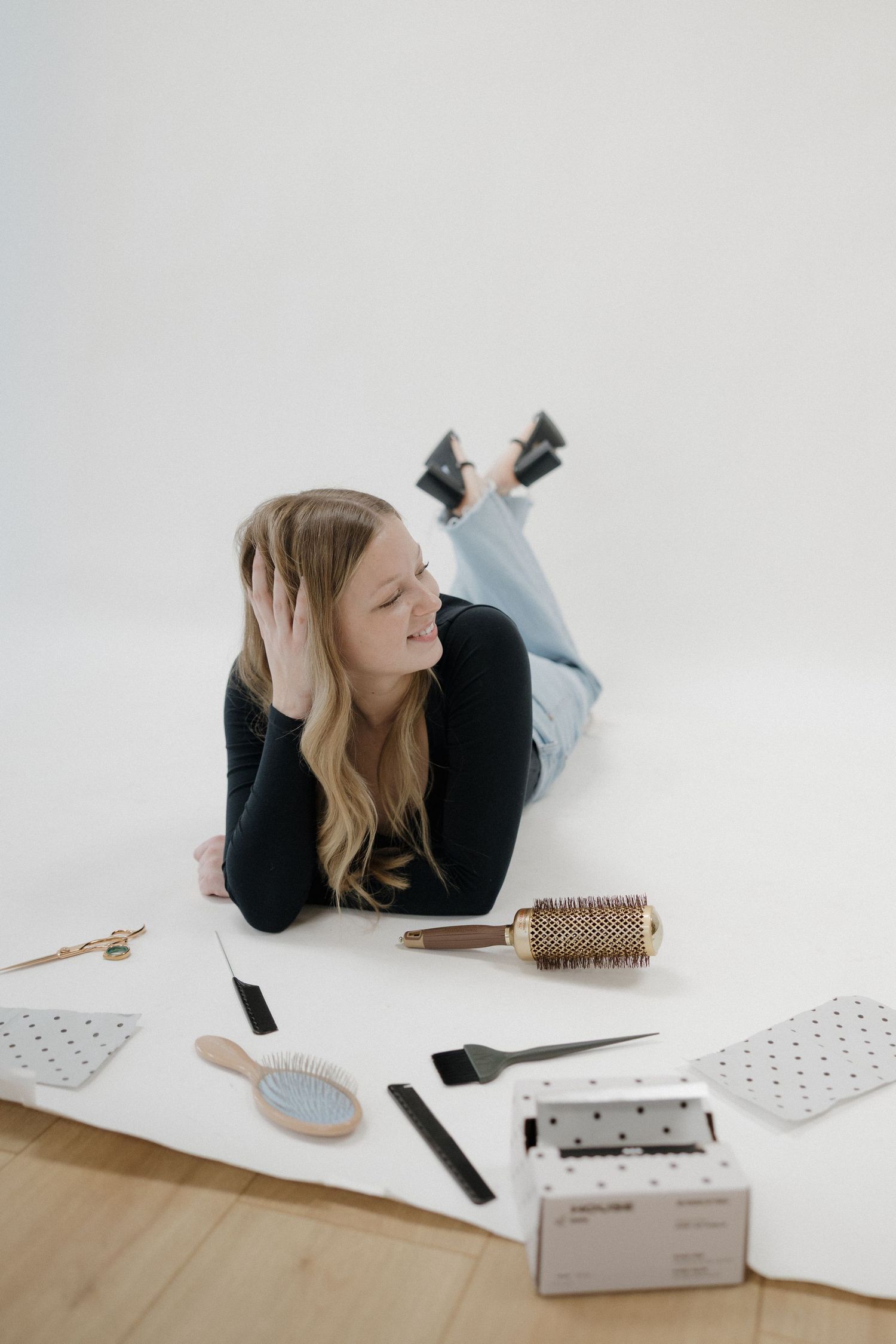 Smiling blonde woman lying on white floor surrounded by professional hair styling tools and brushes.