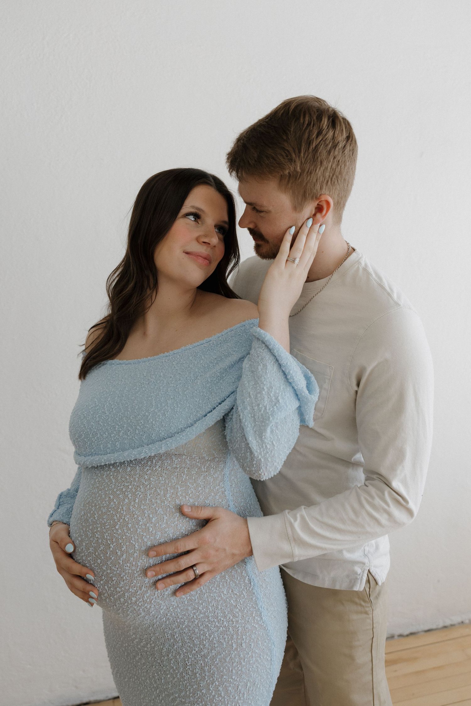 Pregnant couple embracing tenderly, woman in blue off-shoulder dress cradling baby bump, man in cream shirt gazing lovingly.