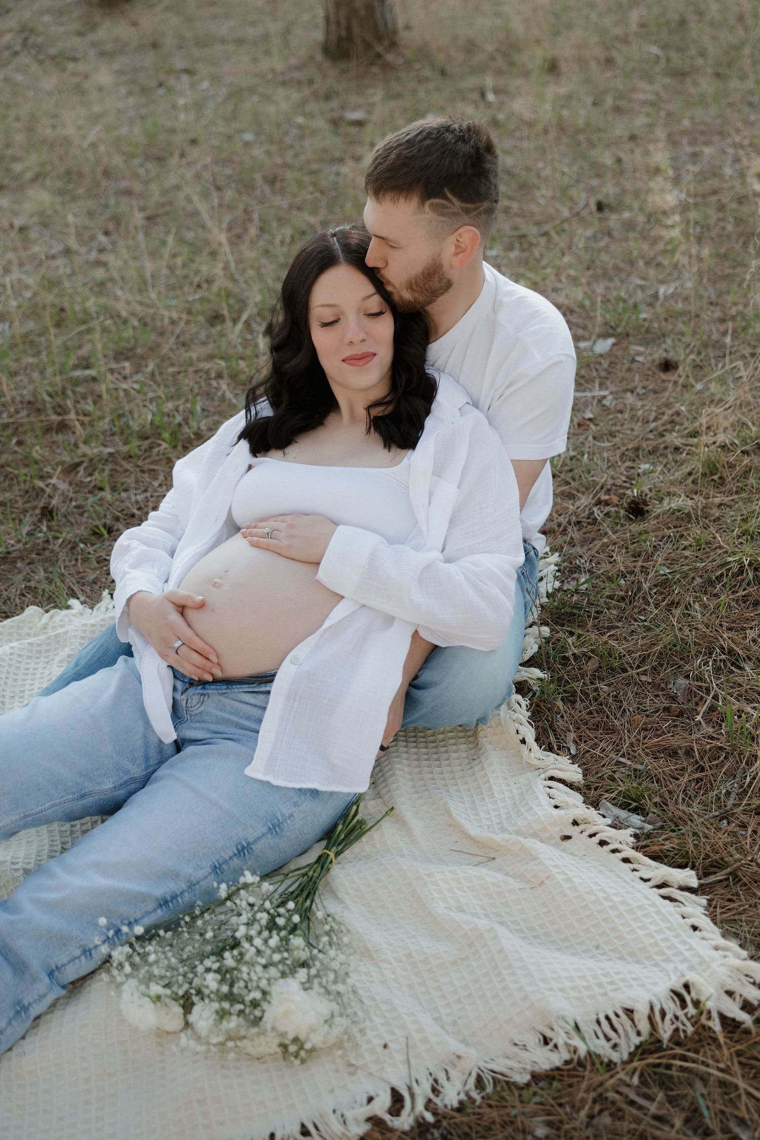 Pregnant couple sitting on white blanket in forest, man kissing woman's head while holding her bare bump.