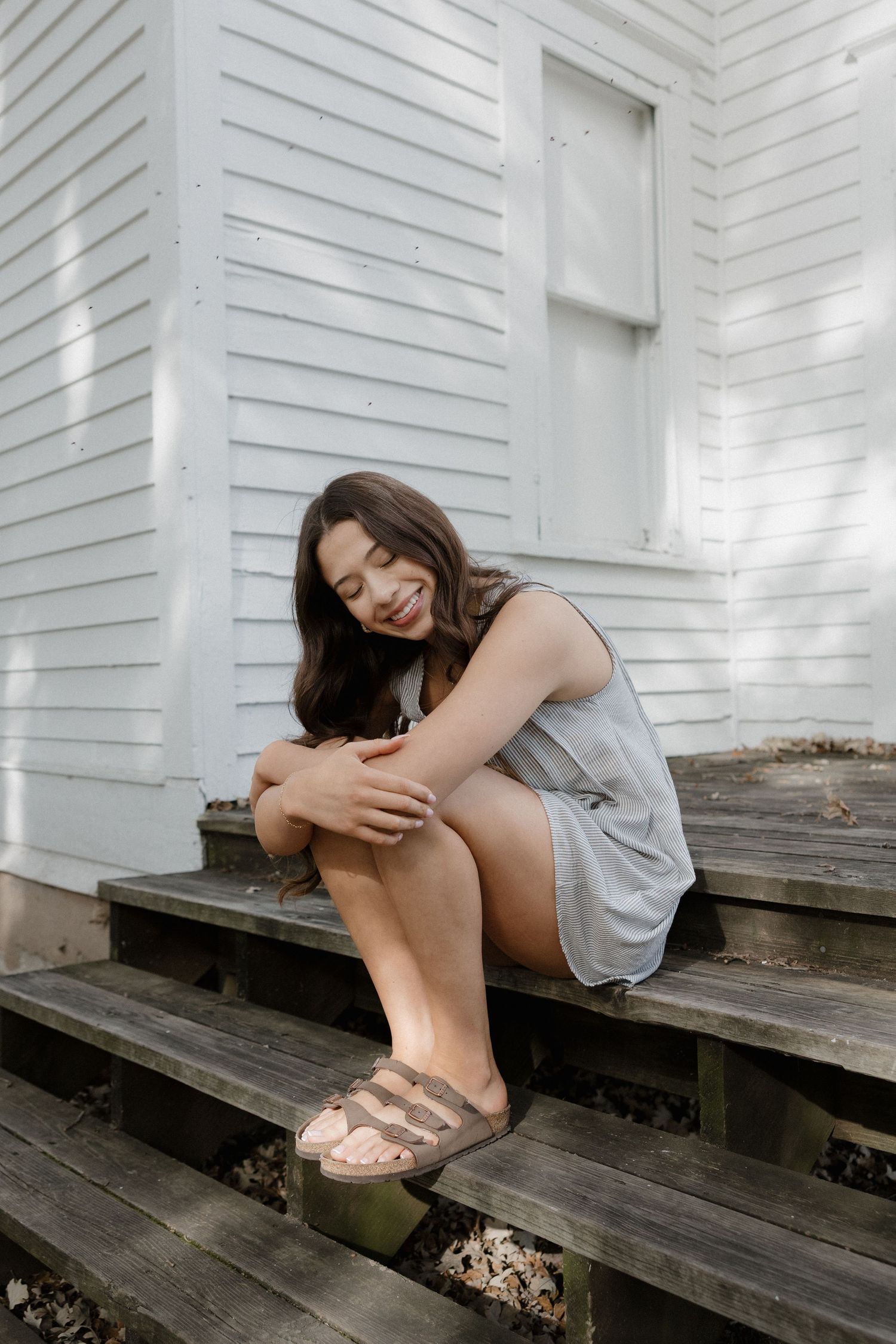 Young woman in gray romper and sandals sitting on wooden steps smiling.