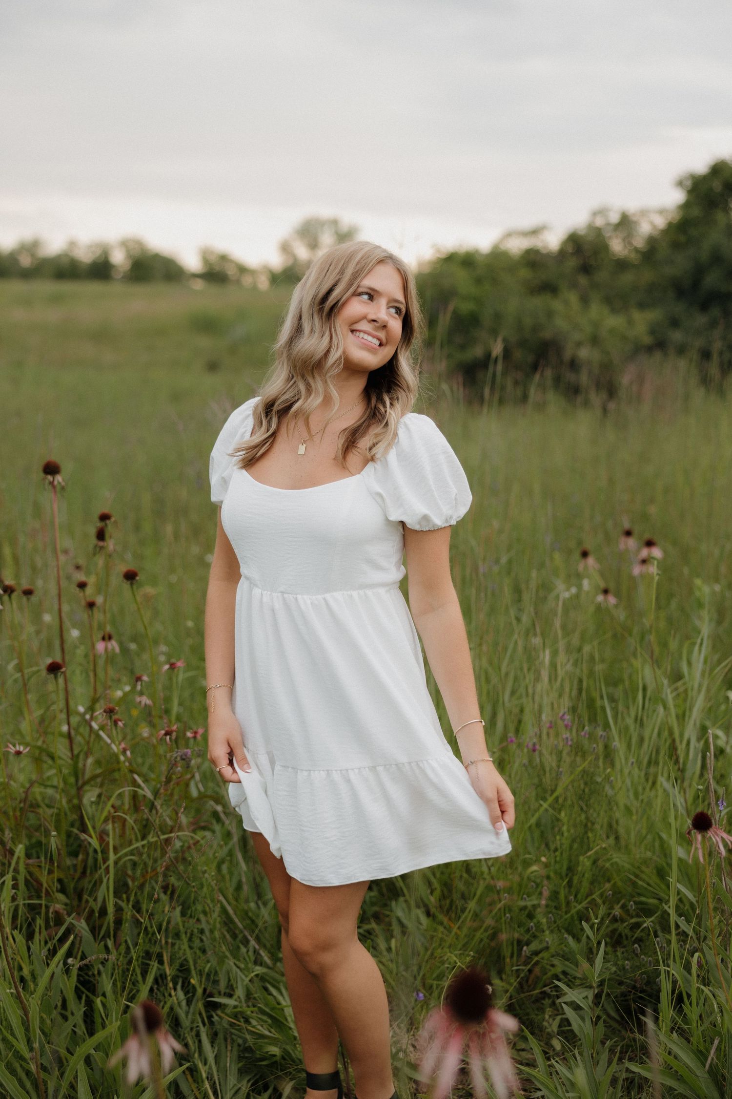Young blonde woman in white puff-sleeve mini dress smiling in a lush wildflower meadow.