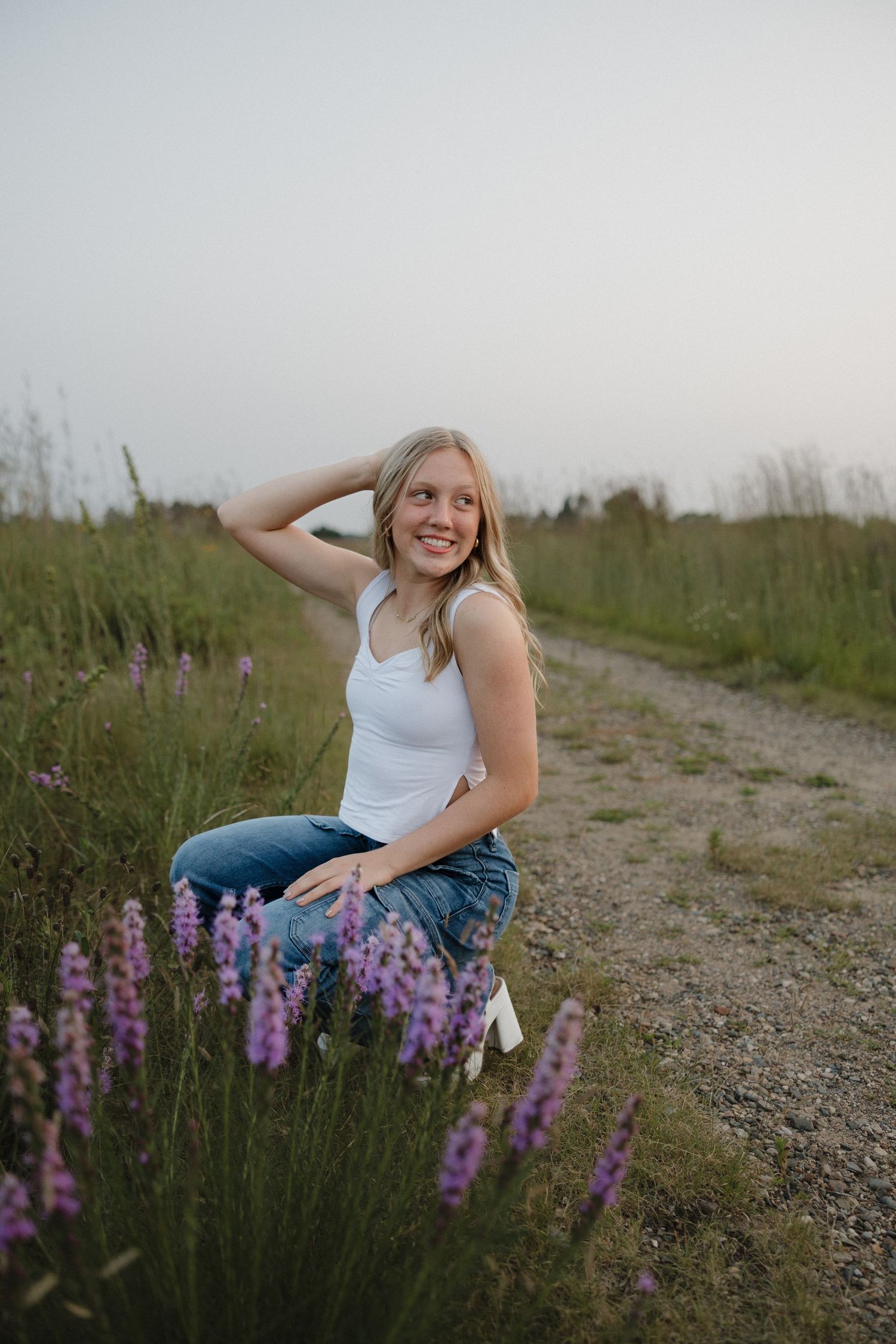 Smiling blonde woman in white tank top and jeans crouching near purple wildflowers on a scenic dirt path.