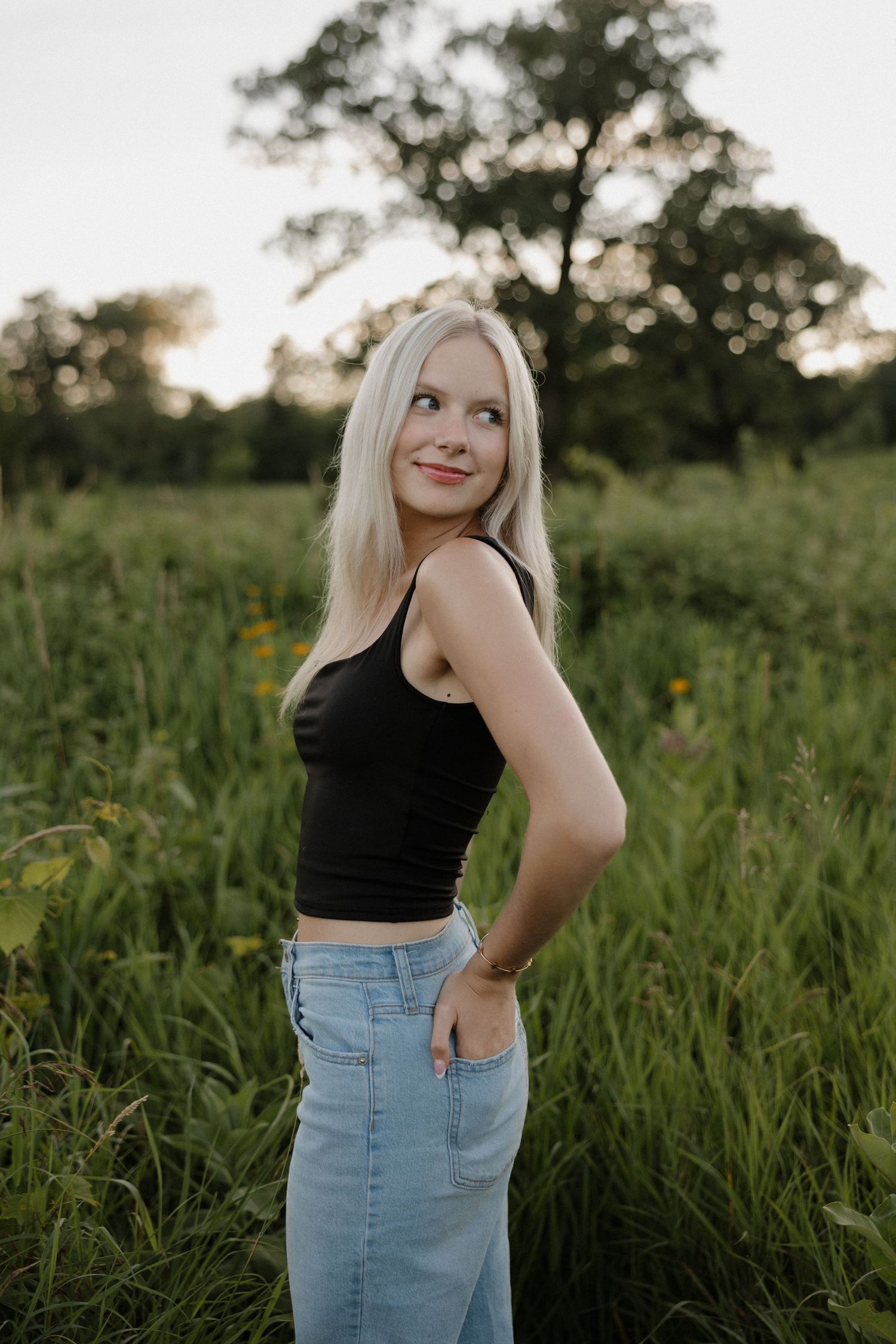 Blonde woman in black crop top and denim skirt posing in a lush green meadow at sunset.