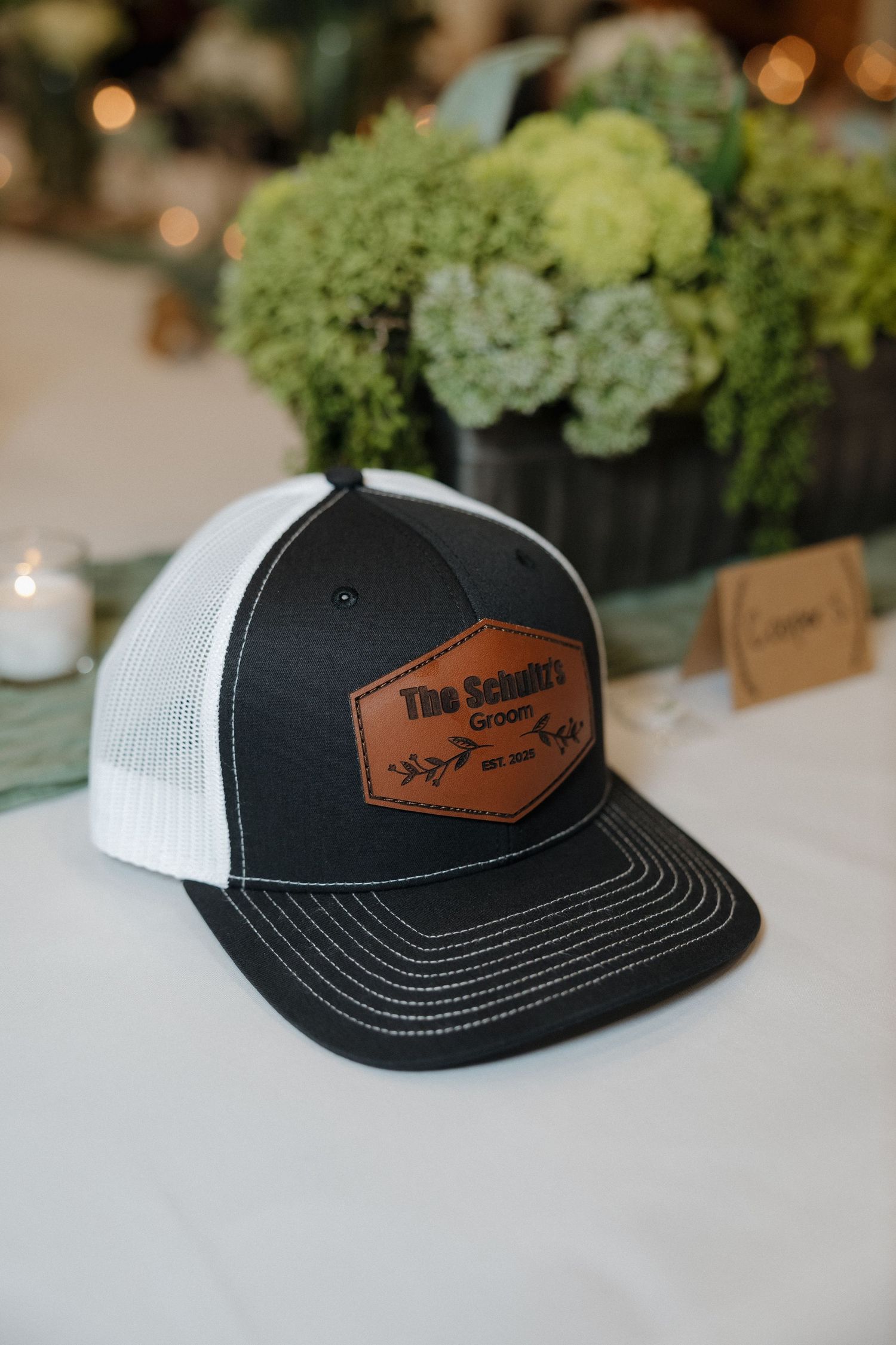 A black and white trucker hat with leather patch sits on a table with green floral arrangements in the background.