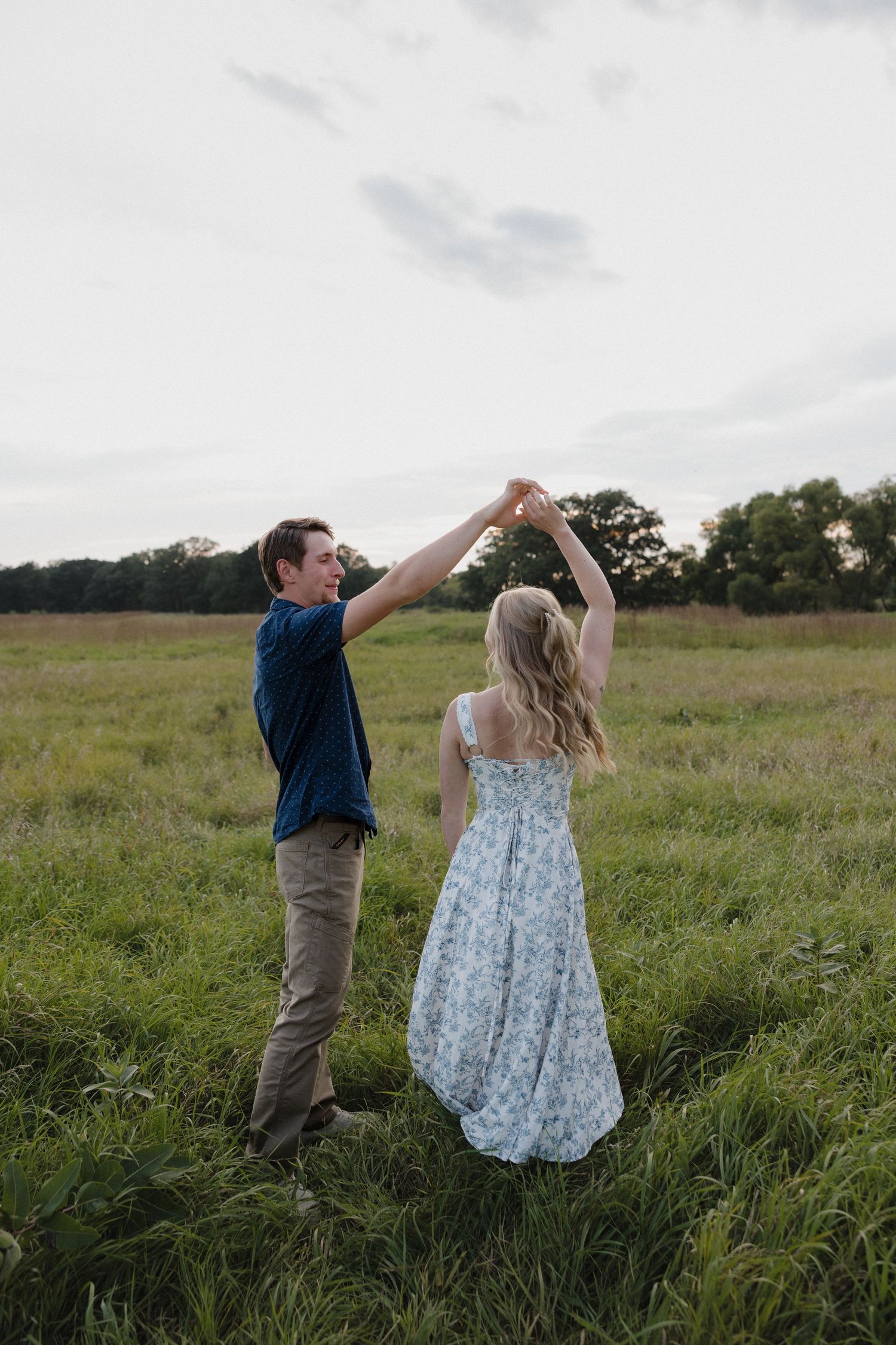 Couple dancing in a lush green field, man spinning woman in floral dress at sunset.
