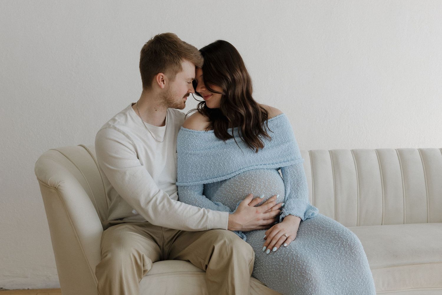 Couple sitting on cream sofa, man tenderly touching pregnant womans blue knit dress belly.