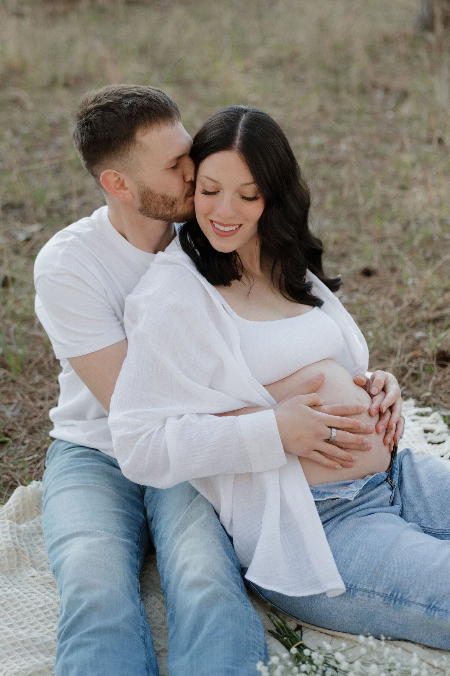 Couple sitting outdoors on blanket, man kissing pregnant woman's cheek while embracing her baby bump.