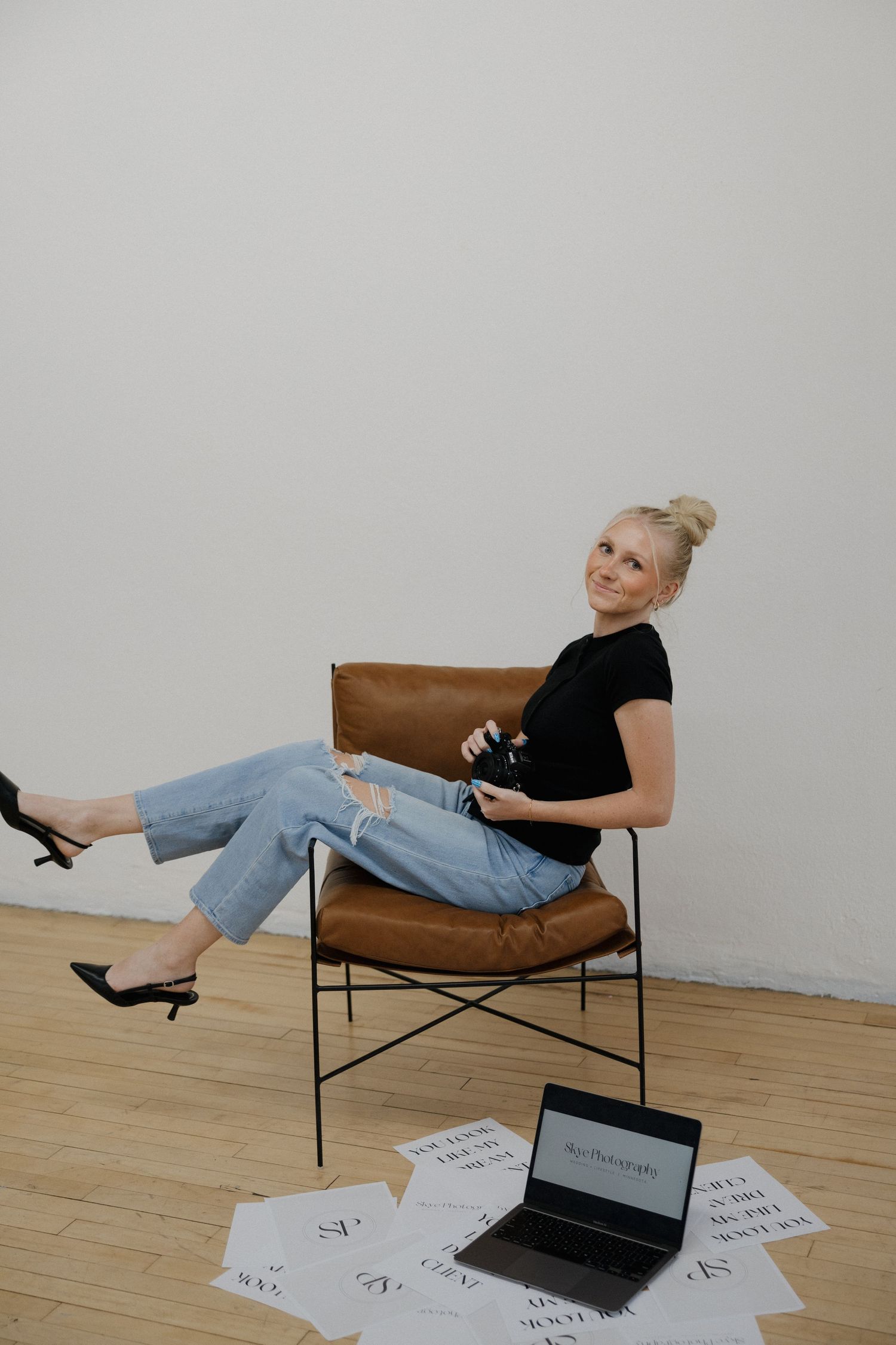 Smiling blonde photographer relaxing in brown leather chair with camera and branded prints.