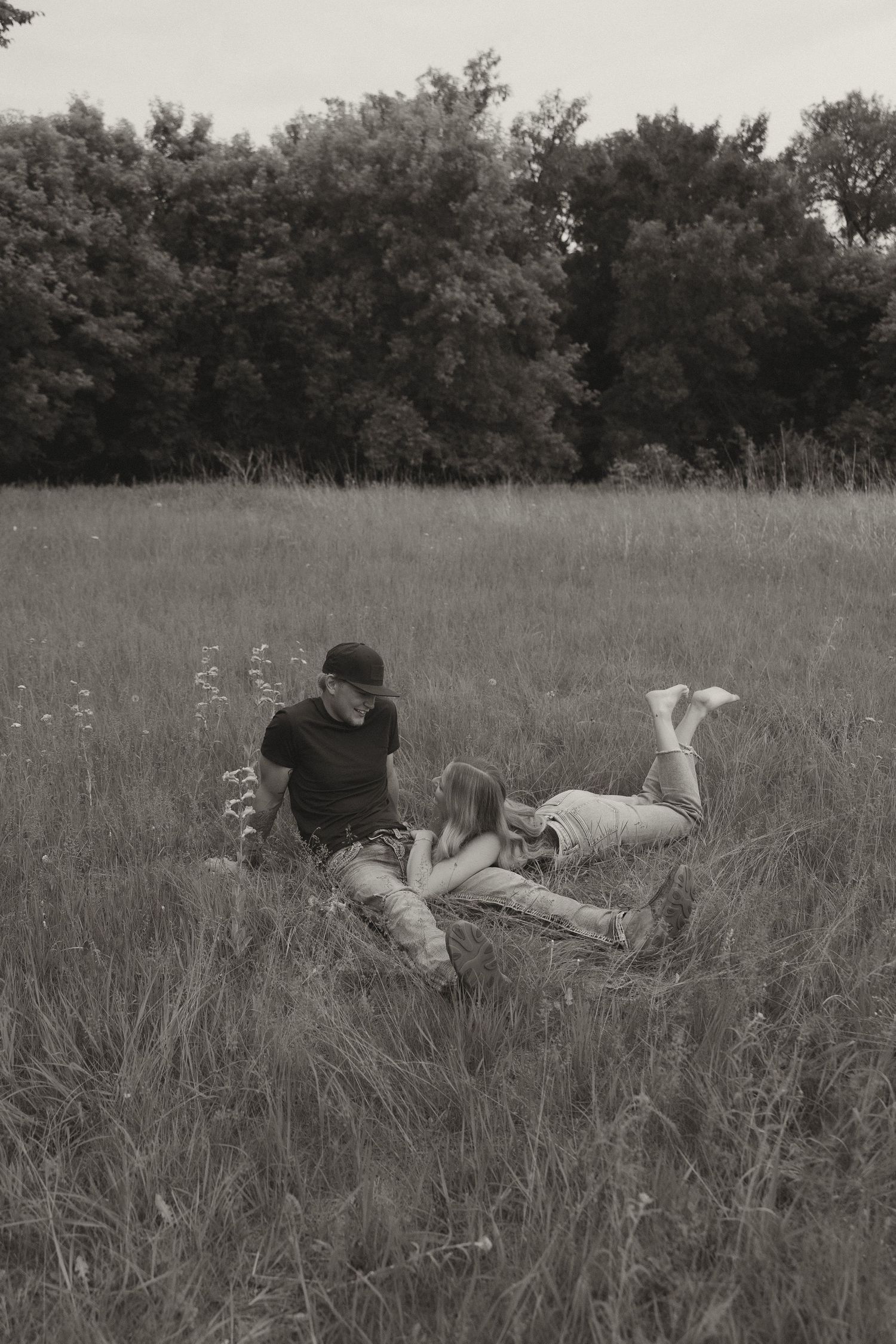 Black and white photo of couple relaxing together in tall grass field surrounded by trees.