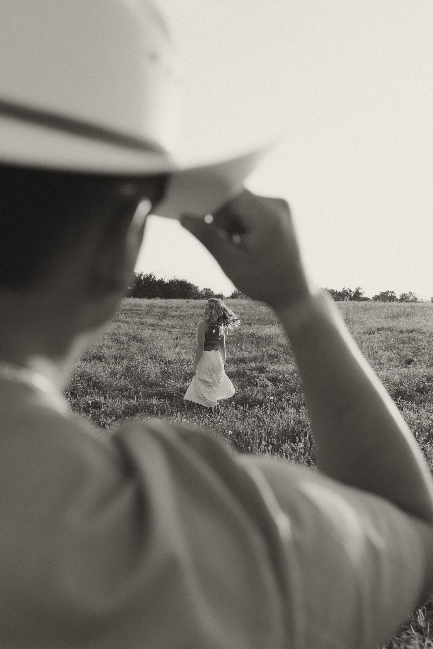 Black and white photo of man adjusting white hat while woman stands in distant field holding flowers.