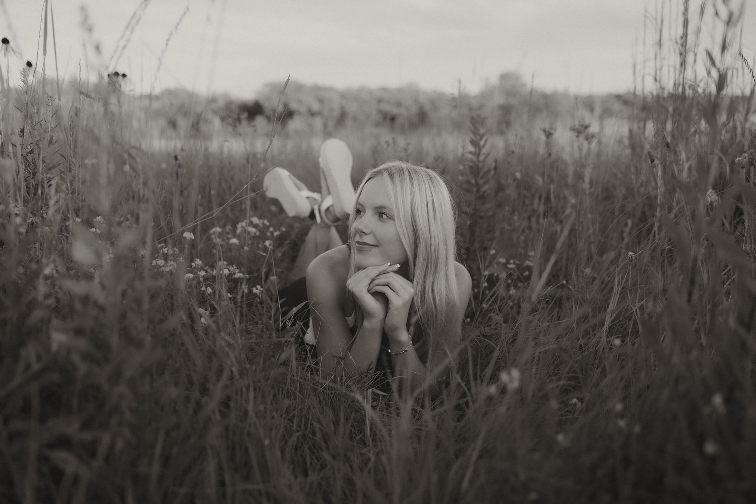 Black and white photo of a blonde woman lying in tall grass holding a wildflower.